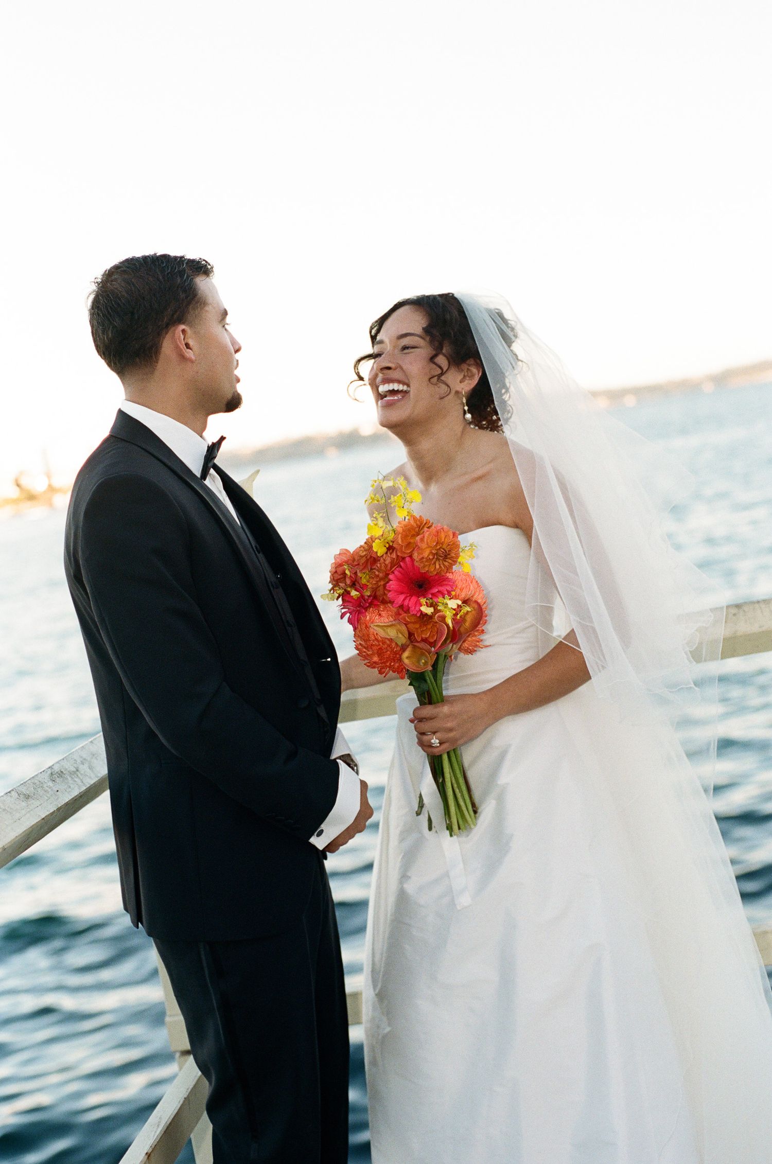 Smiling bride in a white gown and veil holds an orange bouquet while laughing with the groom in a tuxedo by the waterfront.