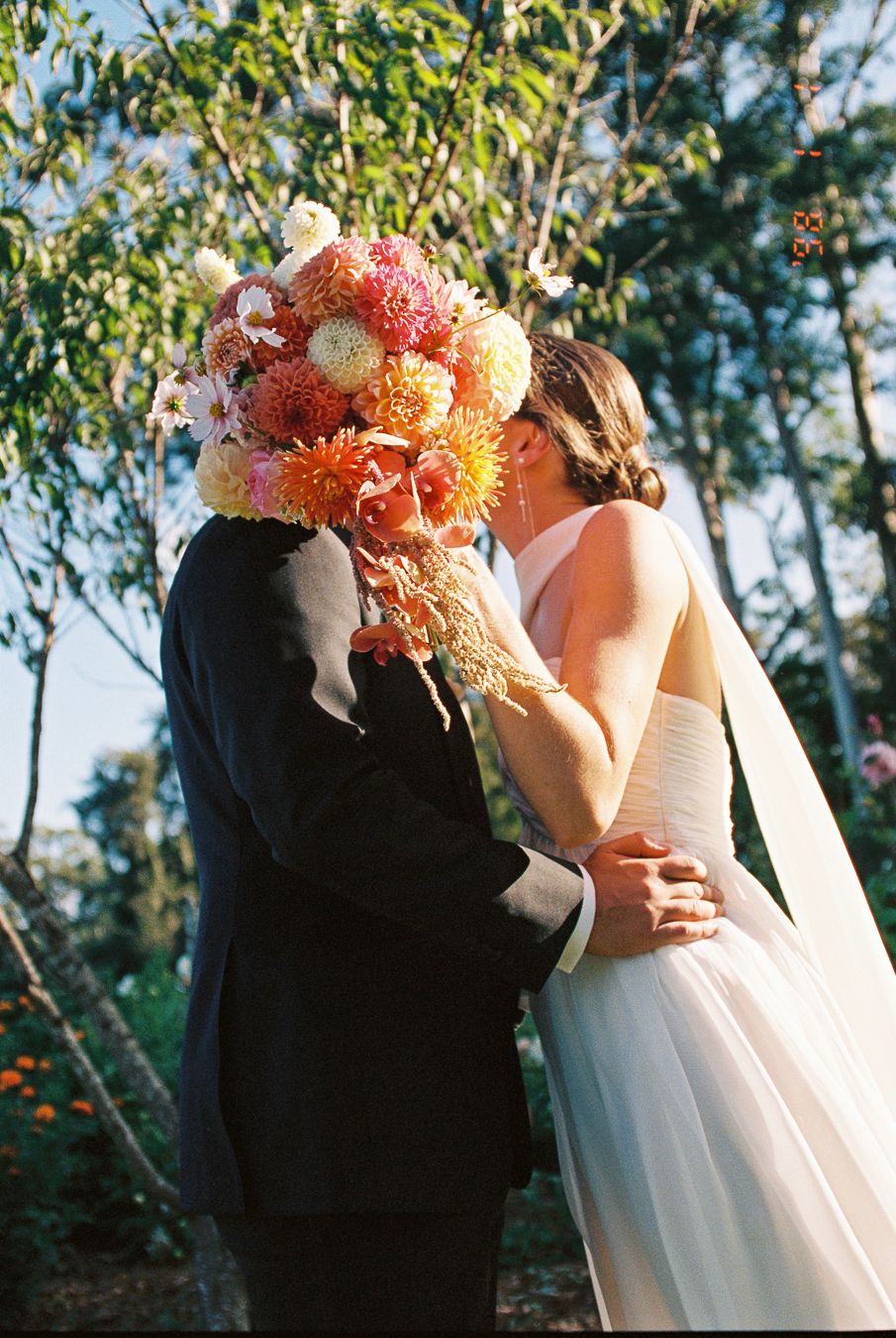 Bride and groom kissing behind a vibrant bouquet of pink and orange dahlias at an outdoor wedding ceremony.