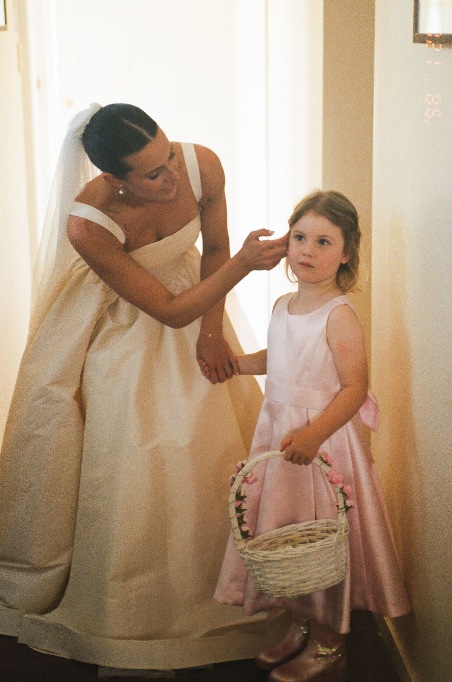 A bride in a strapless ivory gown adjusts a flower girl's hair, as the girl in a pink dress holds a wicker basket of flowers.