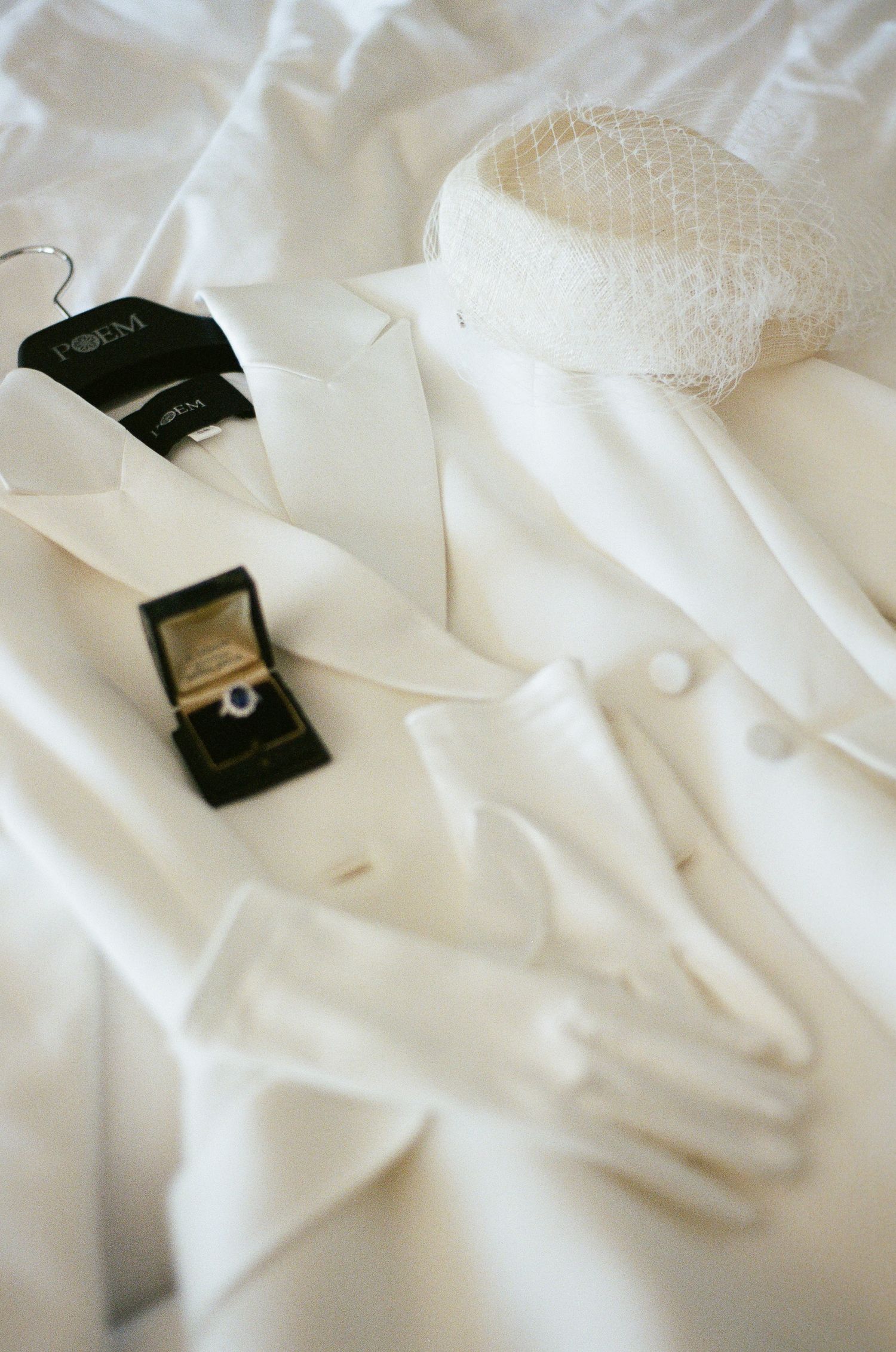 White bridal blazer on a hanger with sheer netted pillbox hat, satin gloves, and an open ring box holding a sapphire engagement ring.