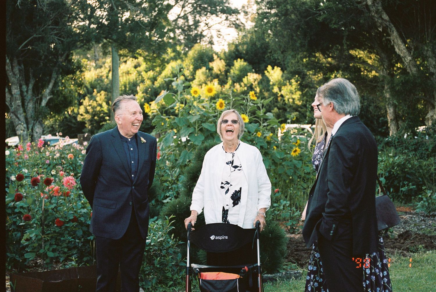 Three people laugh together in a sunny garden, with an elderly woman in white using a walker between two men in dark suits.