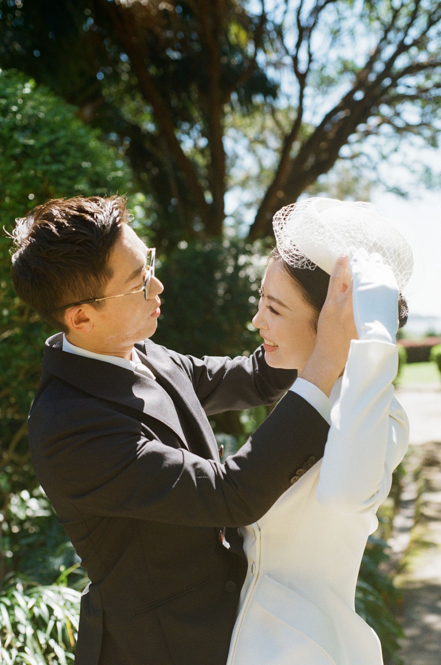 Groom in dark suit and glasses gently adjusts the white lace veil of his smiling bride in a white coat, set against sunlit green trees.