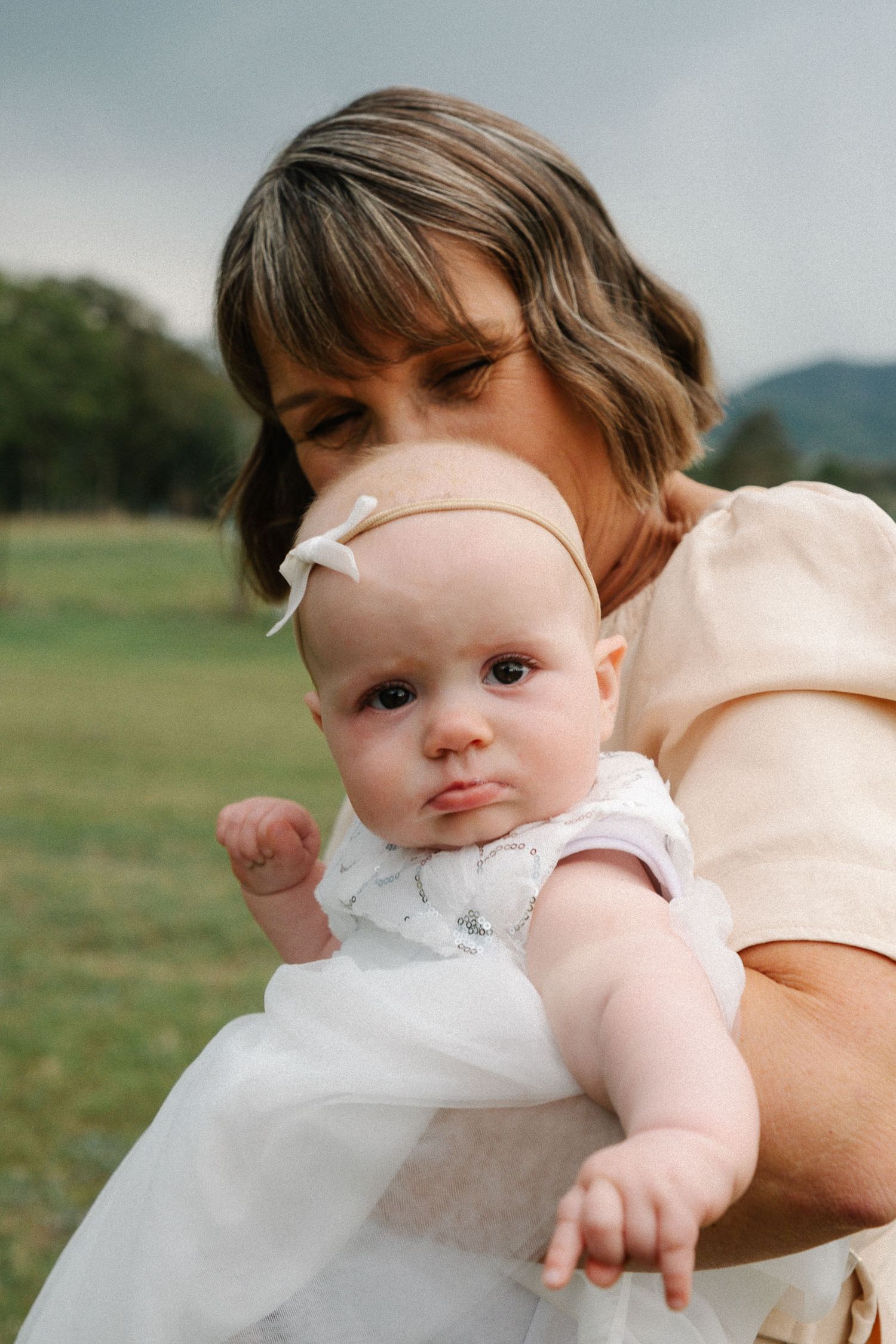 A baby in white clothing is being held while laying down outdoors in a grassy setting.