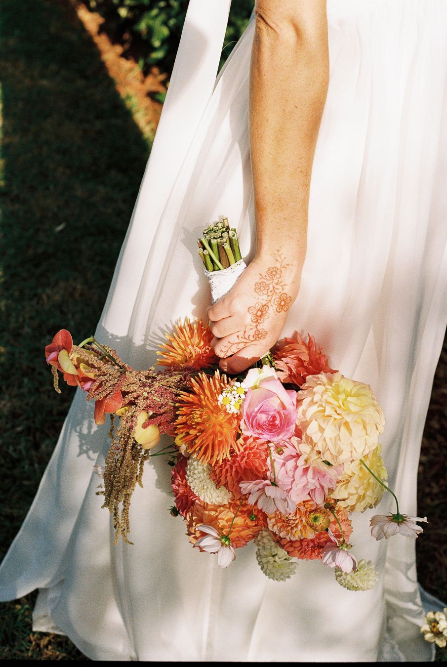 Bride in white gown holding colorful wedding bouquet with dahlias and roses, henna-decorated hand, outdoor setting.