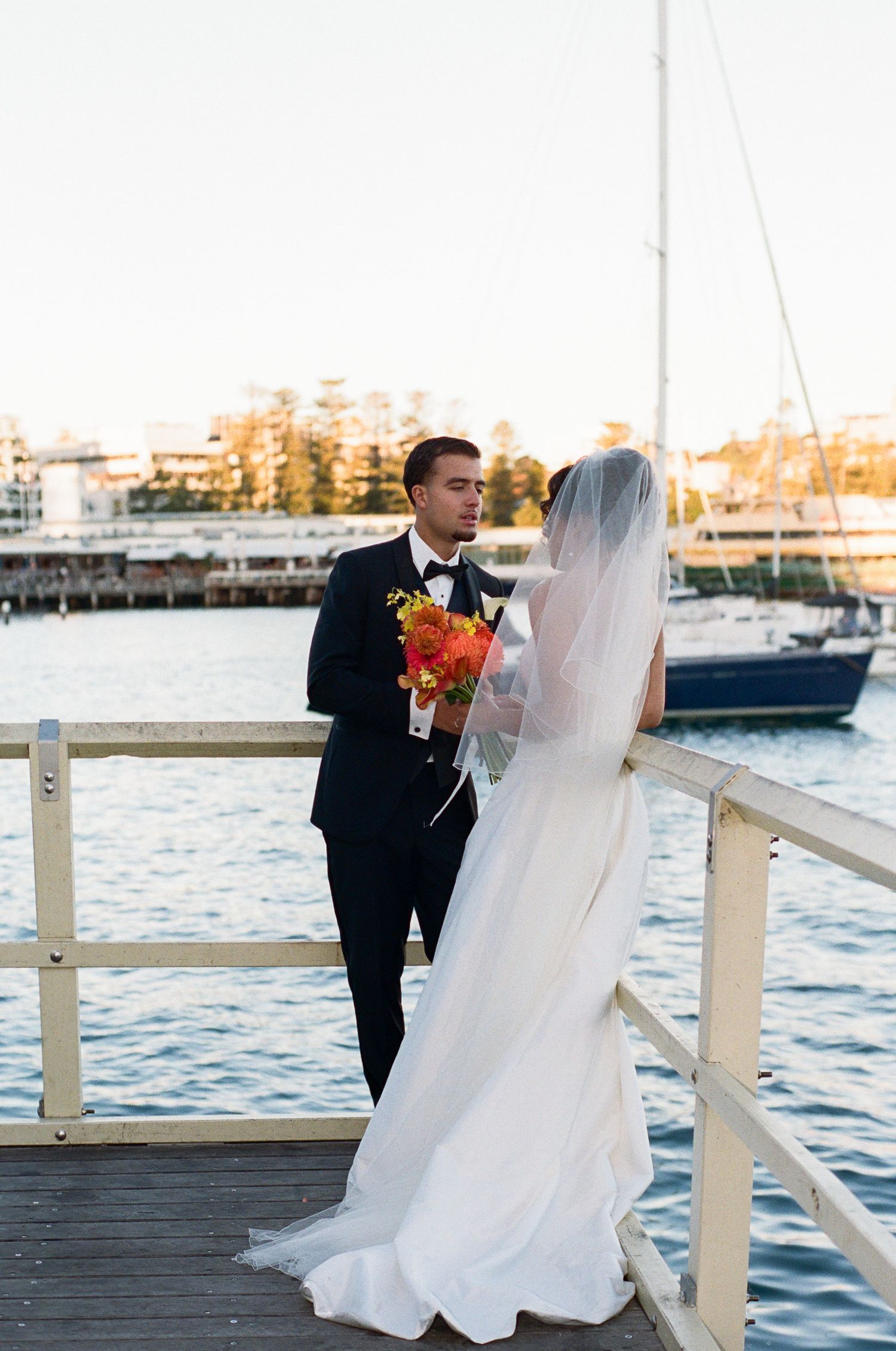 Bride in a flowing white gown and veil faces a groom in a black tuxedo holding an orange bouquet on a waterfront pier with sailboats.