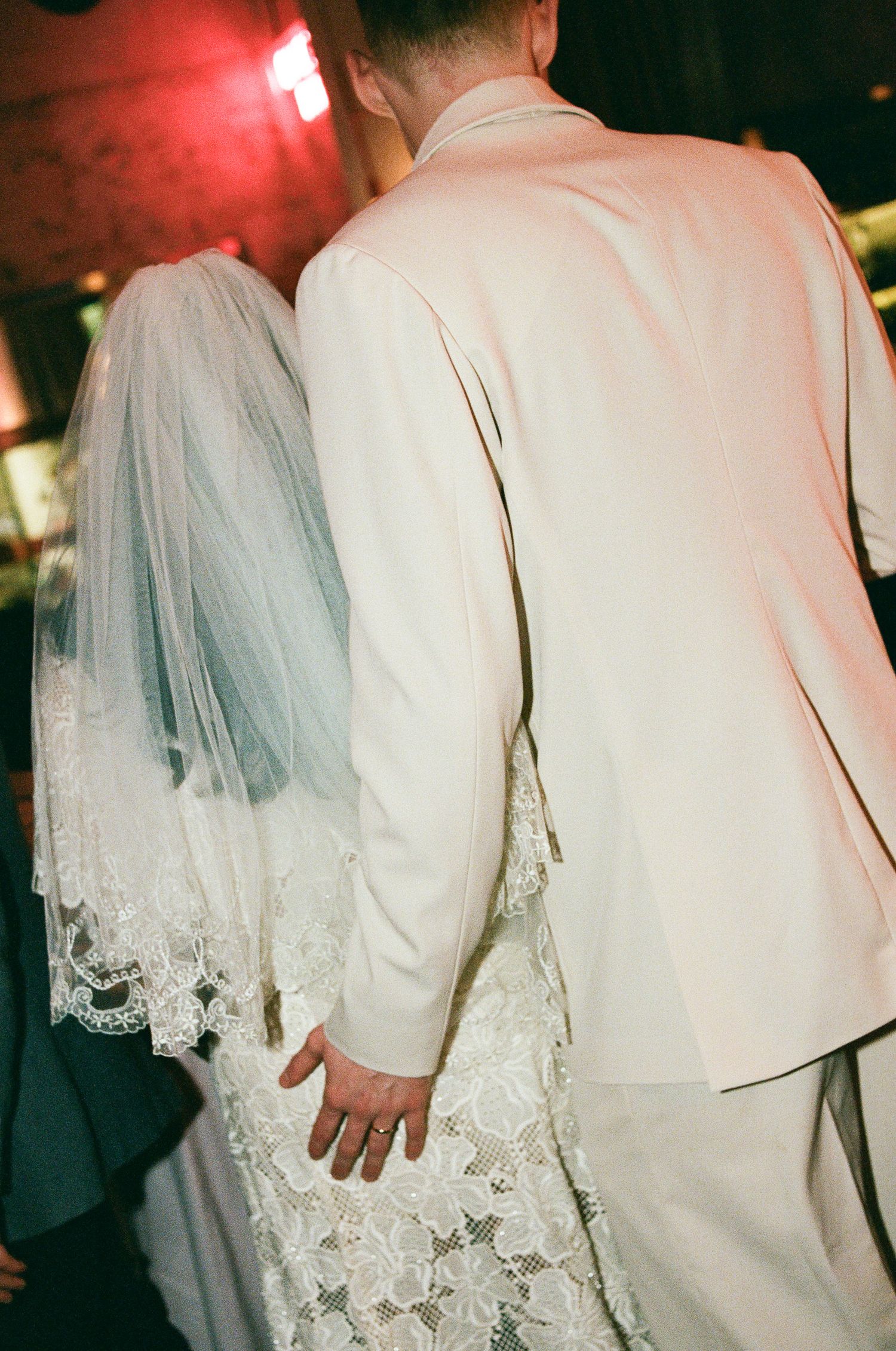 Bride in a lace wedding dress and sheer veil stands beside a groom in a cream suit jacket, seen from behind at a wedding reception.
