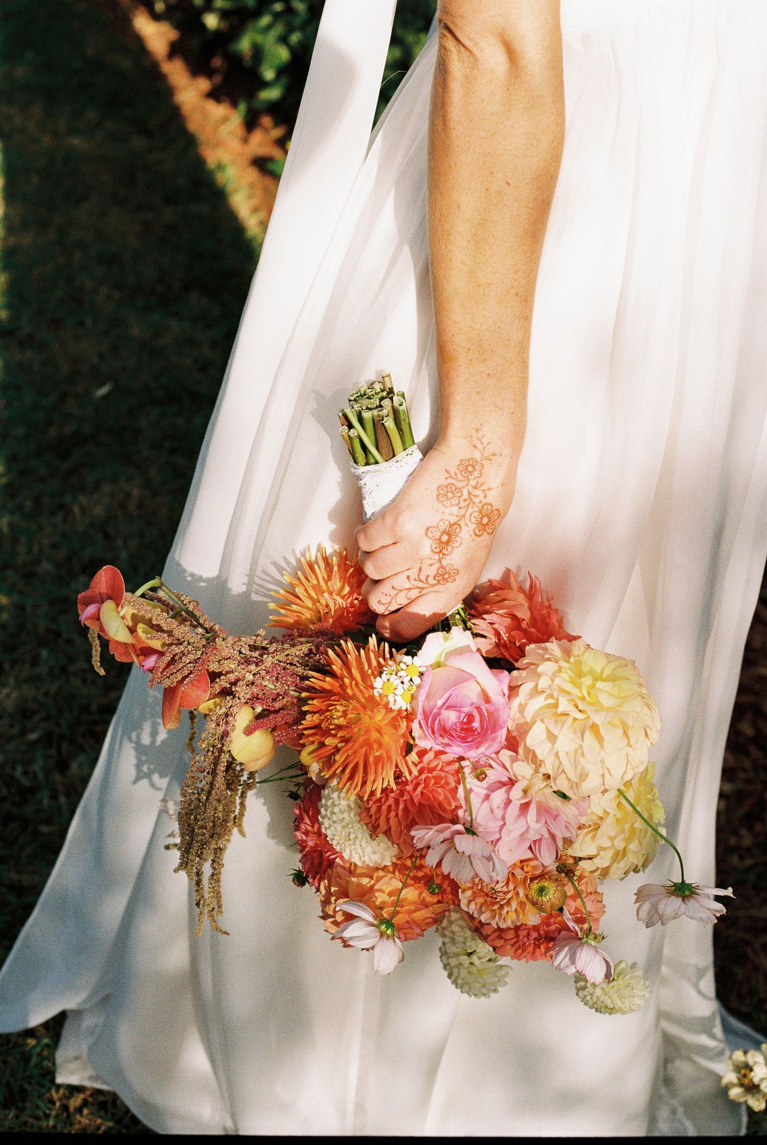 Bride in a flowing white gown holds a colorful dahlia bouquet in coral, pink and orange, her hand decorated with delicate henna designs.