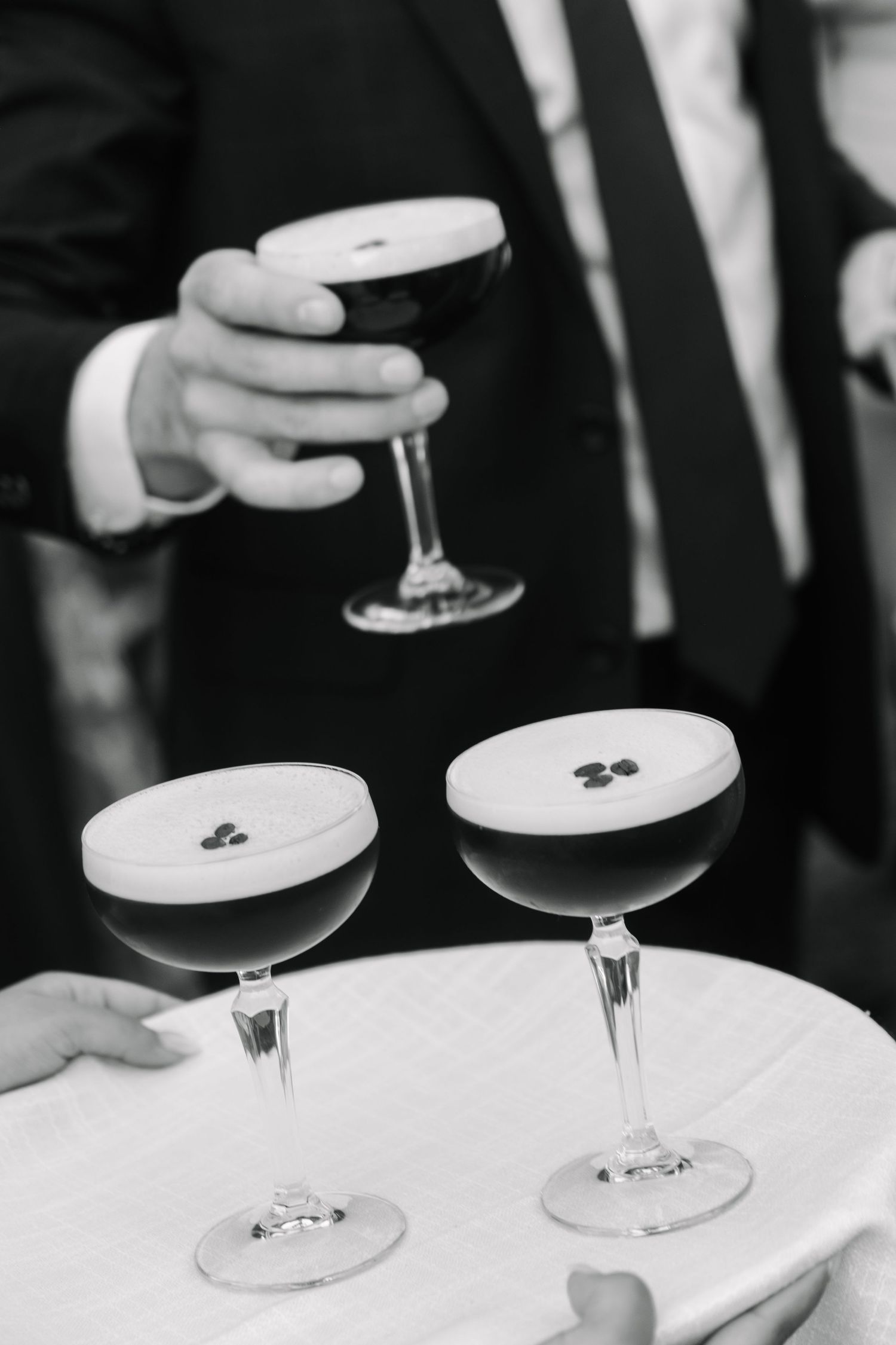 Black and white photo of espresso martini cocktails being served on a tray at a wedding reception.