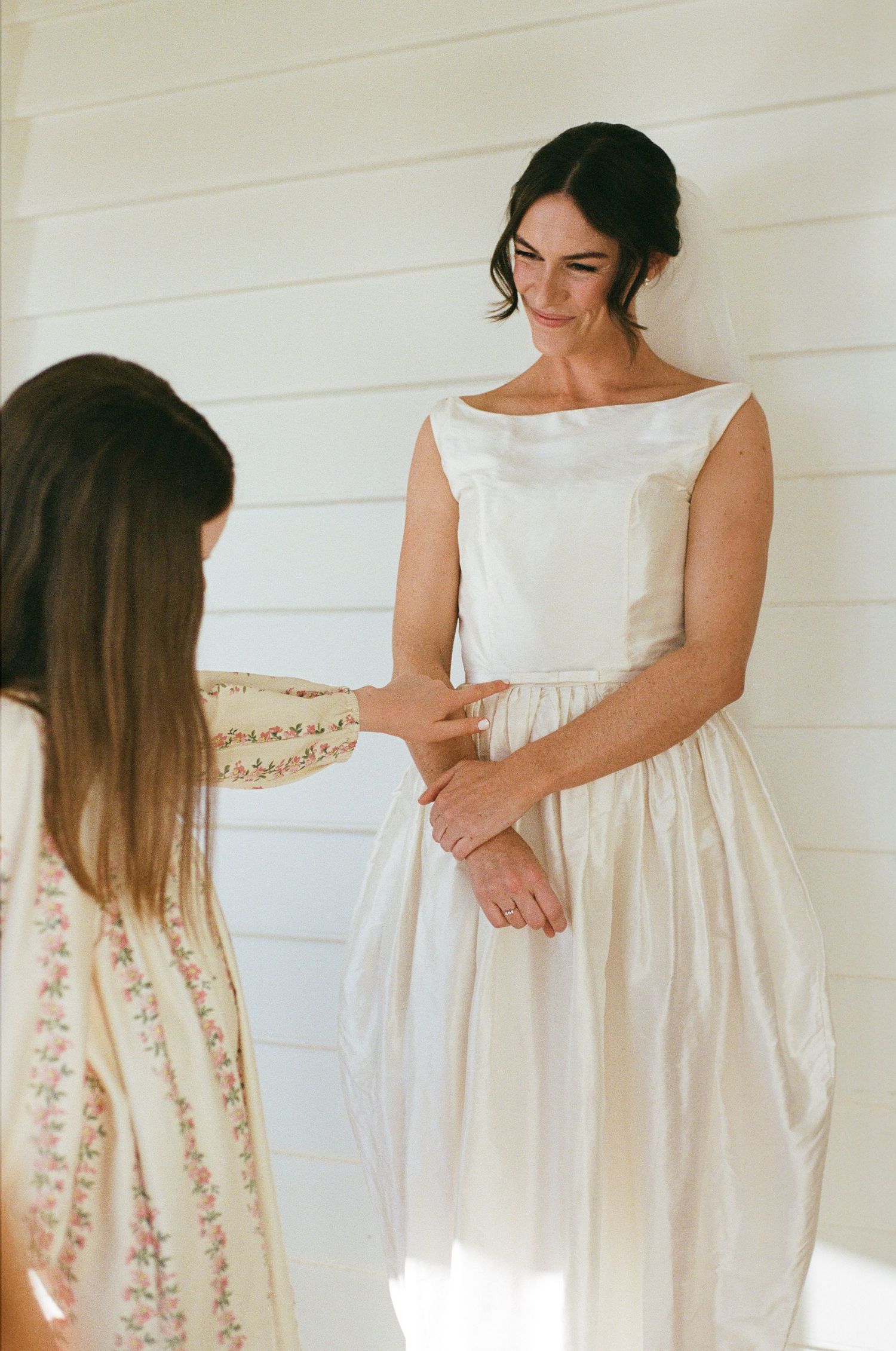 Bride in elegant white satin gown being helped by bridesmaid in floral dress against white shiplap wall.