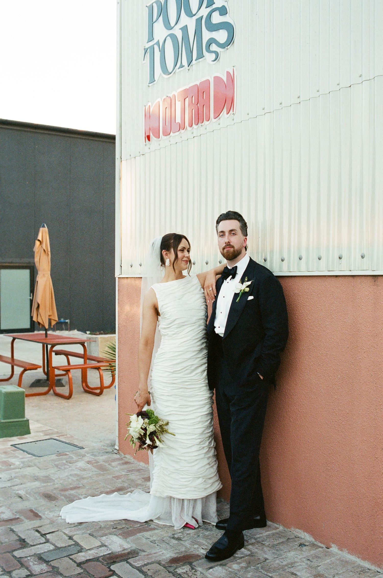 Bride in a ruched white wedding gown and groom in a black tuxedo pose against a pink and white wall outside Pool Toms with a bouquet.