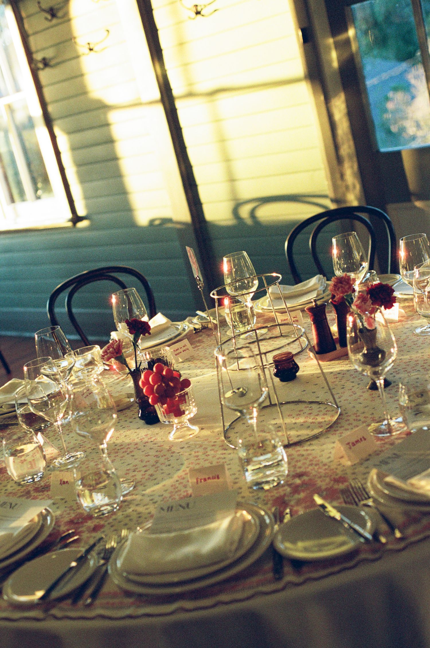 Elegantly set round dining table with white linens, wine glasses, red flower centerpieces, and menu cards in a warmly lit restaurant.