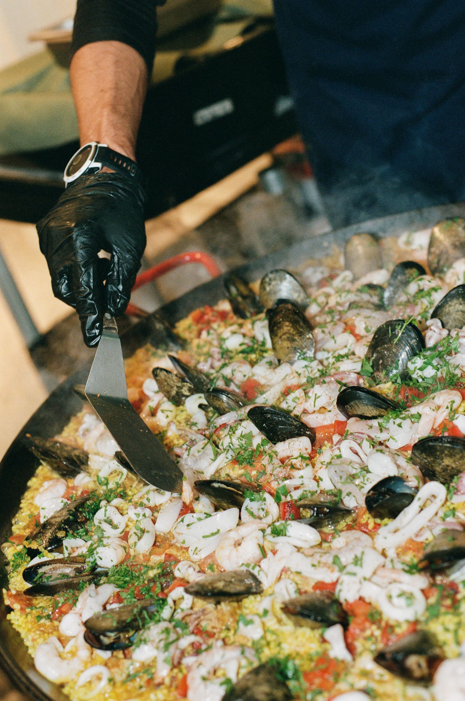 Gloved chef scooping a spatula through a large pan of seafood paella topped with mussels, squid rings, tomatoes and fresh parsley.
