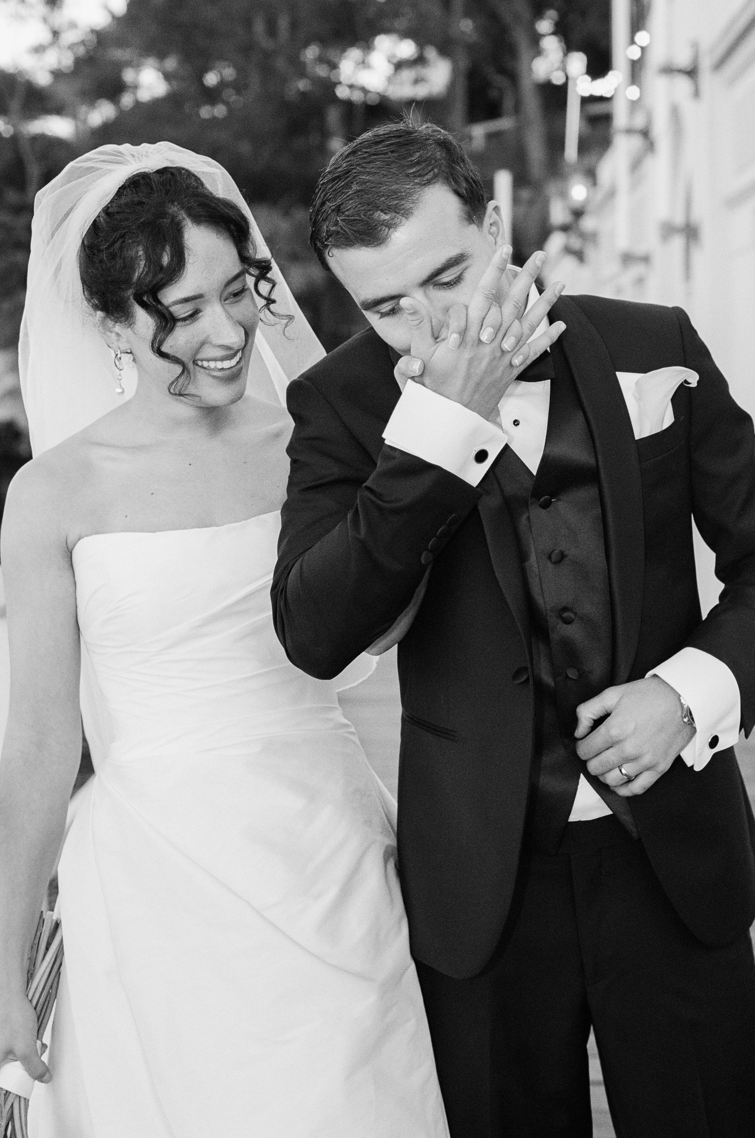 Black and white wedding photo of a smiling bride in a strapless gown as the groom in a tuxedo kisses her hand.
