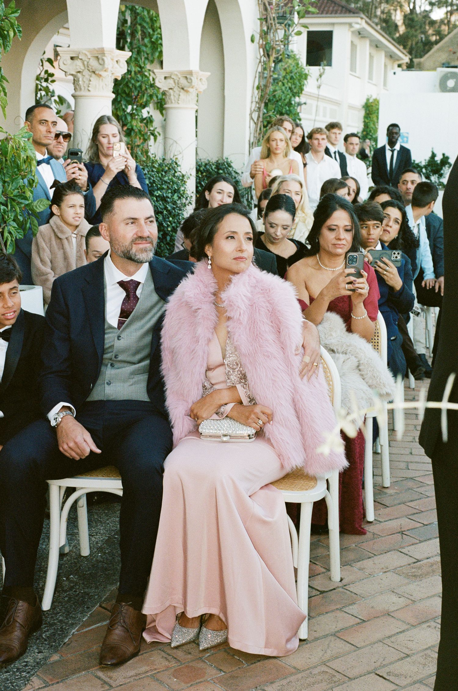 Wedding guests seated on white chairs outdoors, including a bearded man in a navy suit and a woman in a blush gown and pink fur wrap.