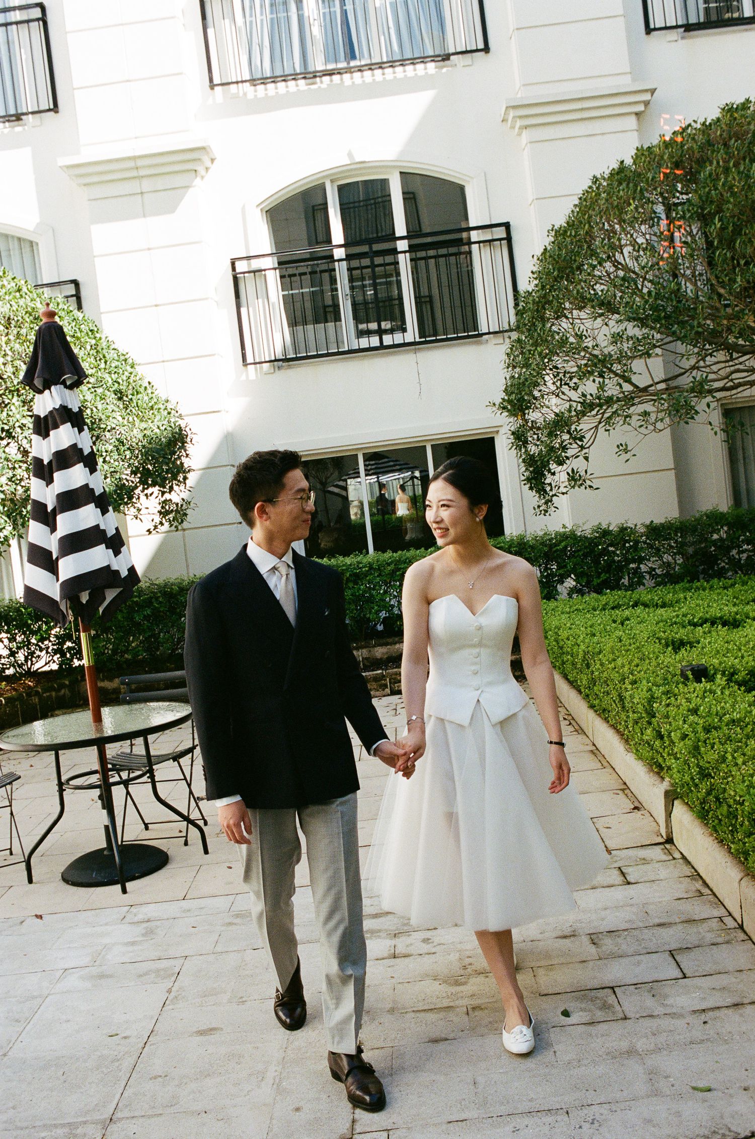 Smiling couple holding hands and walking through a sunlit courtyard, the bride in a white strapless dress and the groom in a dark blazer.