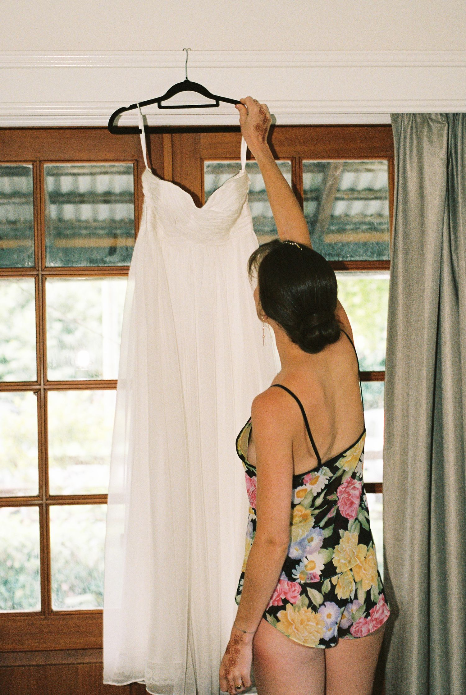 Bride in a floral romper hangs a white strapless wedding gown on a window frame beside sage green curtains on her wedding morning.