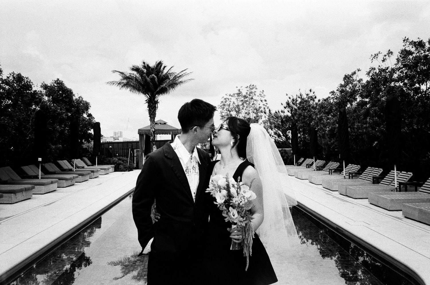 Black and white photo of a bride and groom kissing beside a resort pool lined with lounge chairs and palm trees.