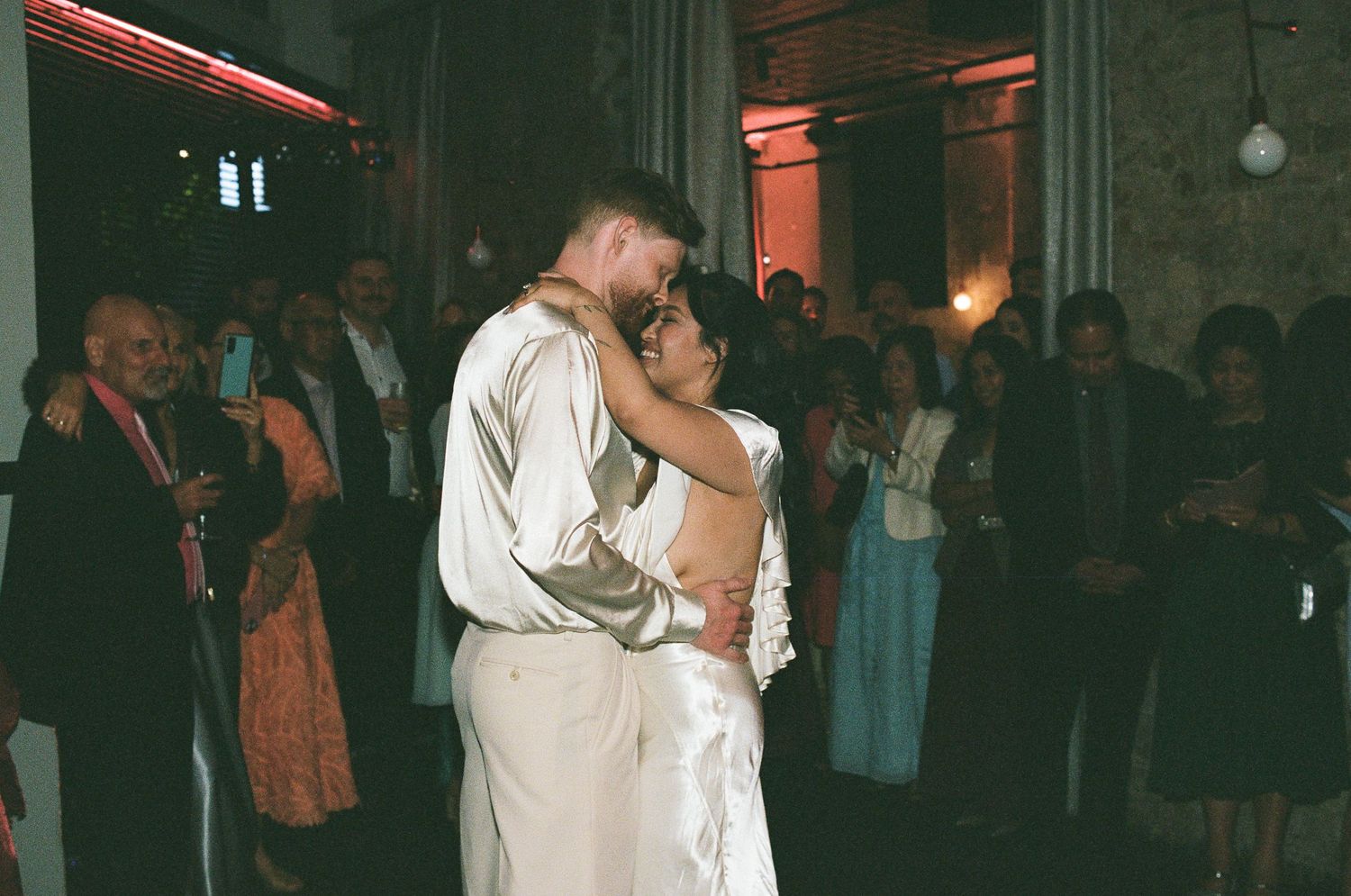 Newlywed couple shares a romantic first dance in a dimly lit reception hall while wedding guests watch and smile in the background.