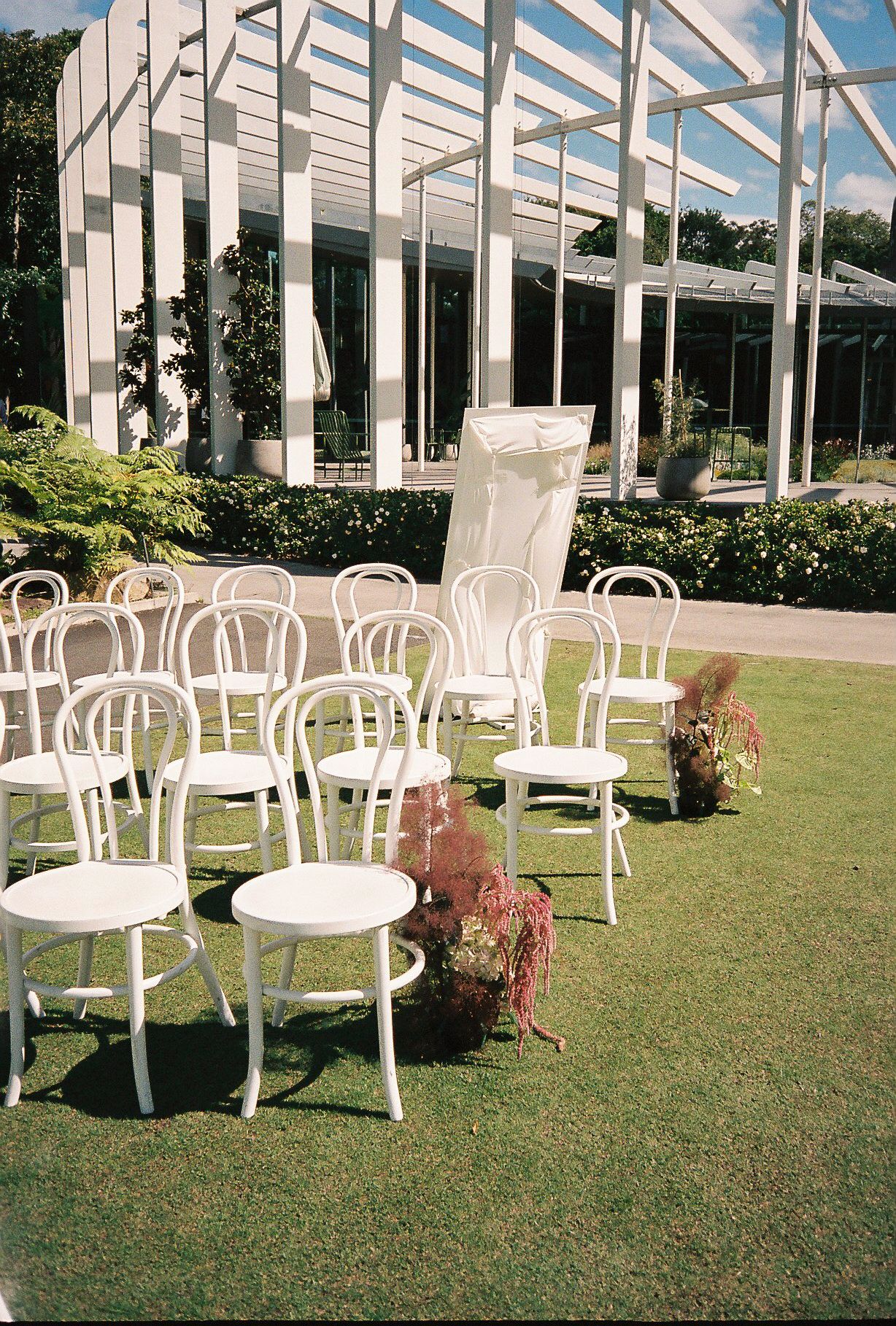 White outdoor chairs arranged on a green lawn under a modern pergola structure with sunlight streaming through.
