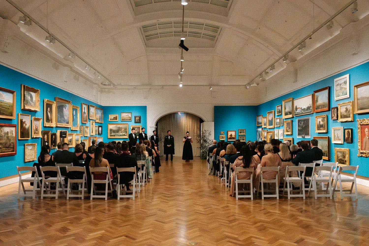 Art gallery visitors view framed artwork displayed on bright turquoise walls in a modern exhibition space.