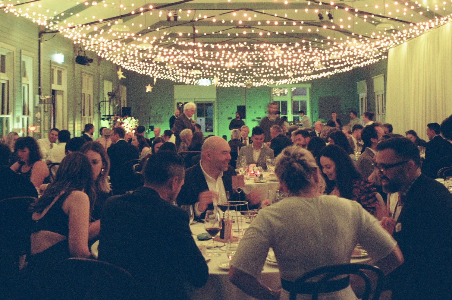 Guests dine at round tables in a warmly lit banquet hall beneath a canopy of string lights during an evening event.