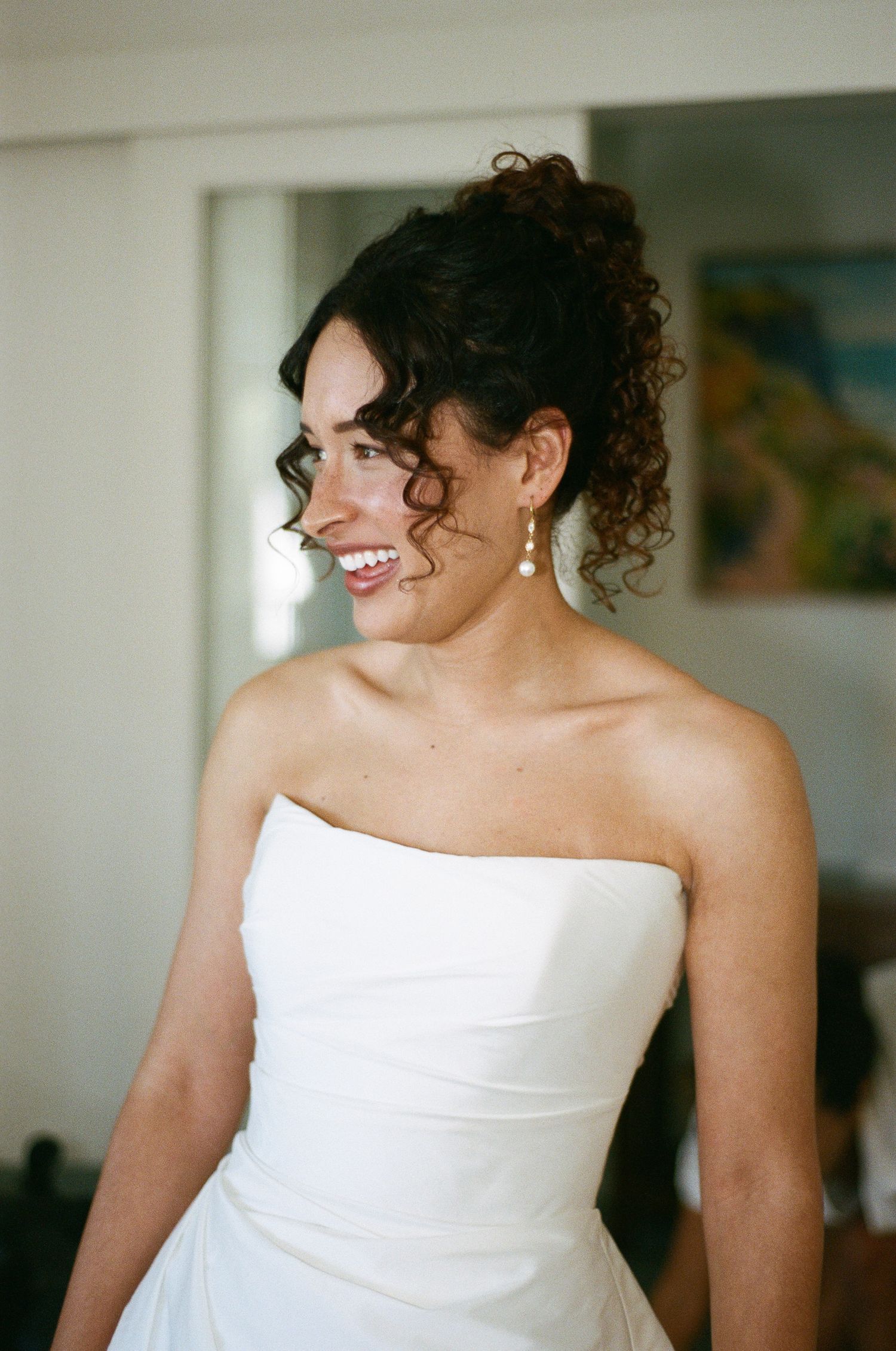 Smiling woman with curly dark hair in an updo wearing a strapless white gown and drop earrings, looking to the side indoors.