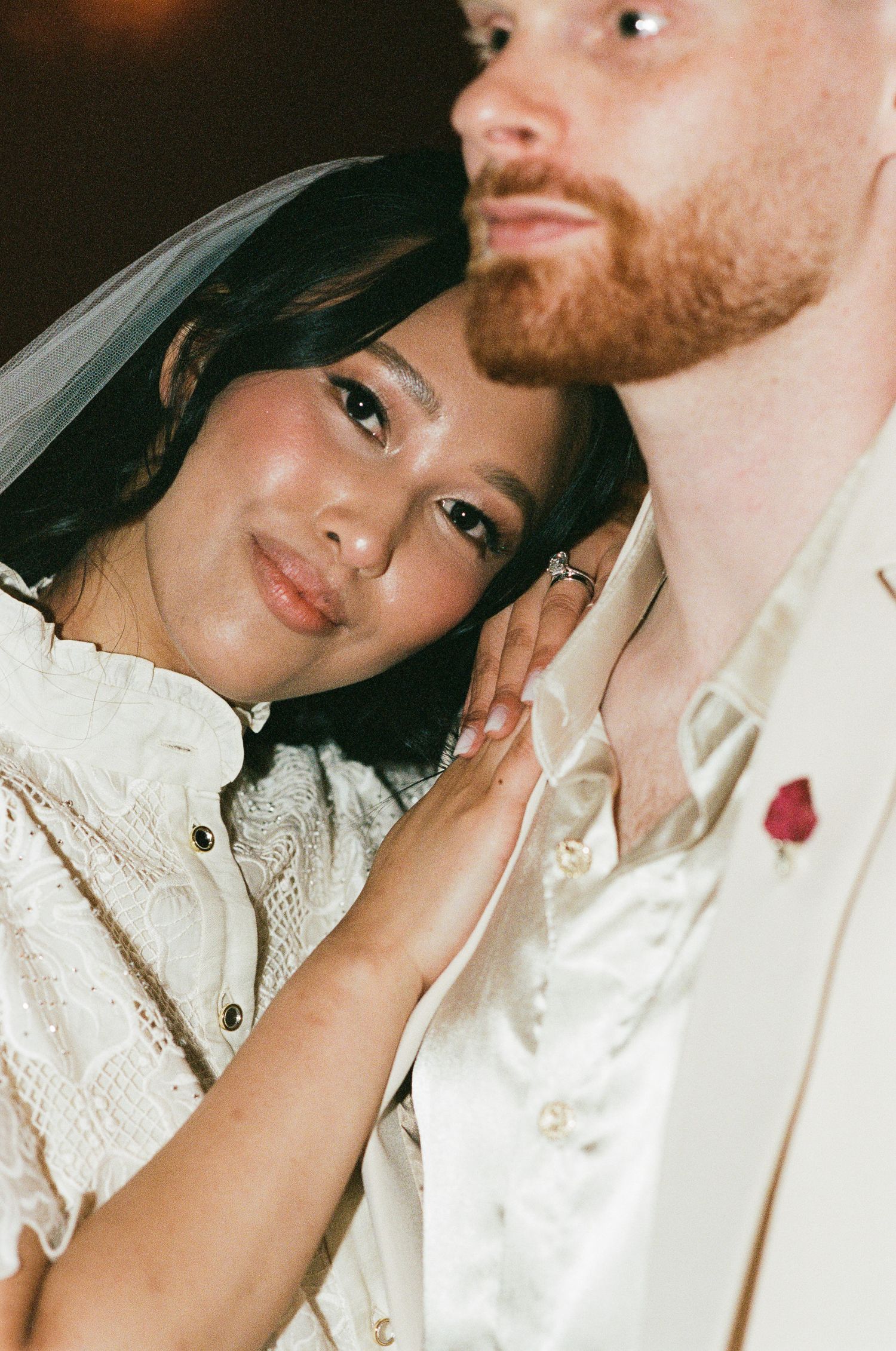 Smiling bride in a lace dress and veil rests her head on her red-bearded groom's shoulder, a red boutonniere on his cream suit.