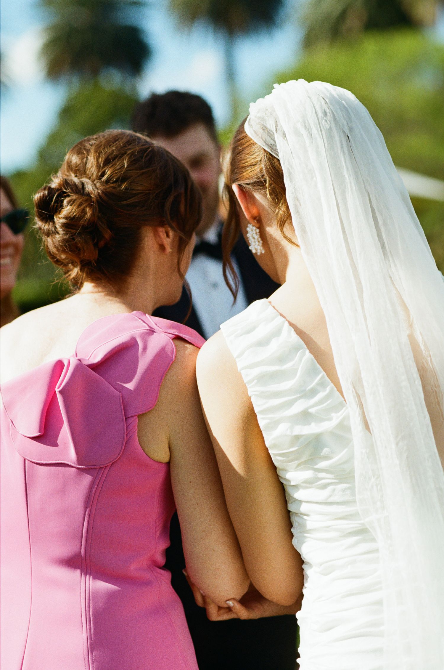 Bride in a white ruched gown and long veil links arms with a woman in a pink dress at an outdoor wedding ceremony.
