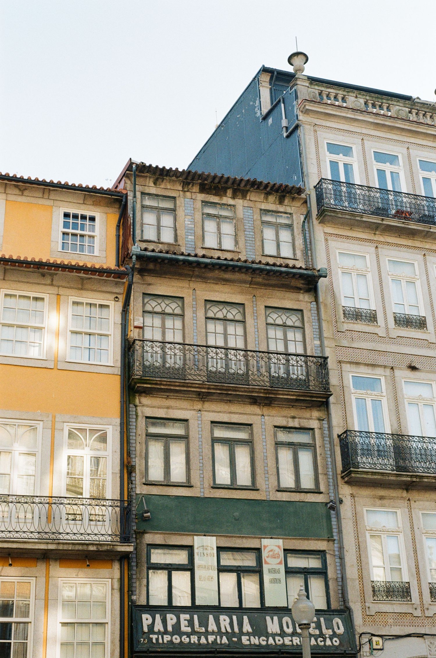 Traditional Portuguese townhouses with wrought-iron balconies and tiled facades above a vintage Papelaria Modelo stationery shop sign.