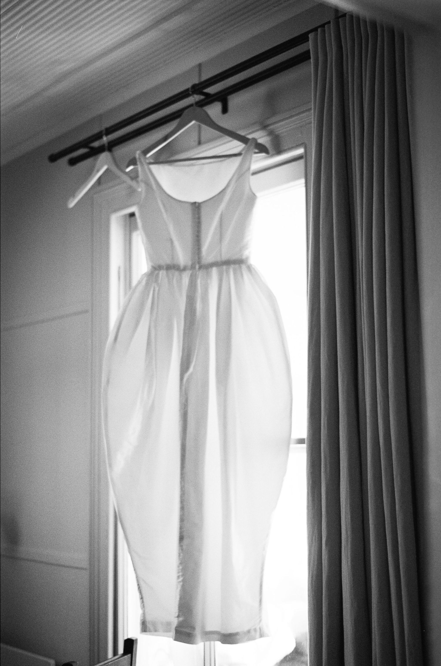 Black and white photo of a white wedding dress hanging on a rod near a window with curtains.
