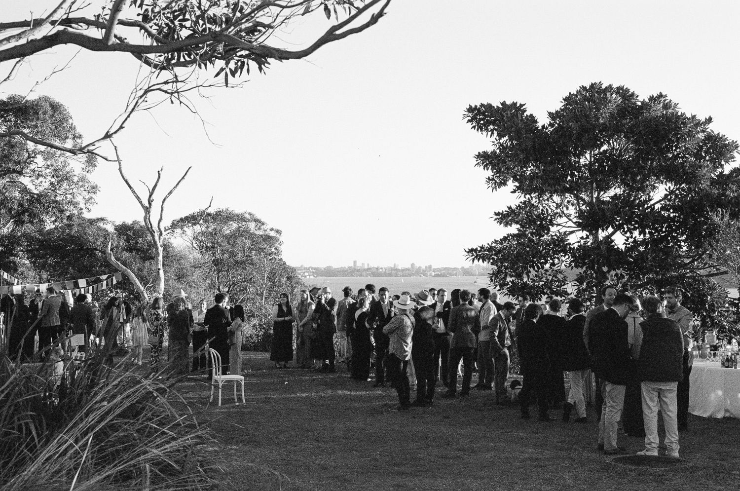 Black and white photo of wedding guests gathered on a grassy lawn beneath trees at an outdoor ceremony with a scenic view in the distance.