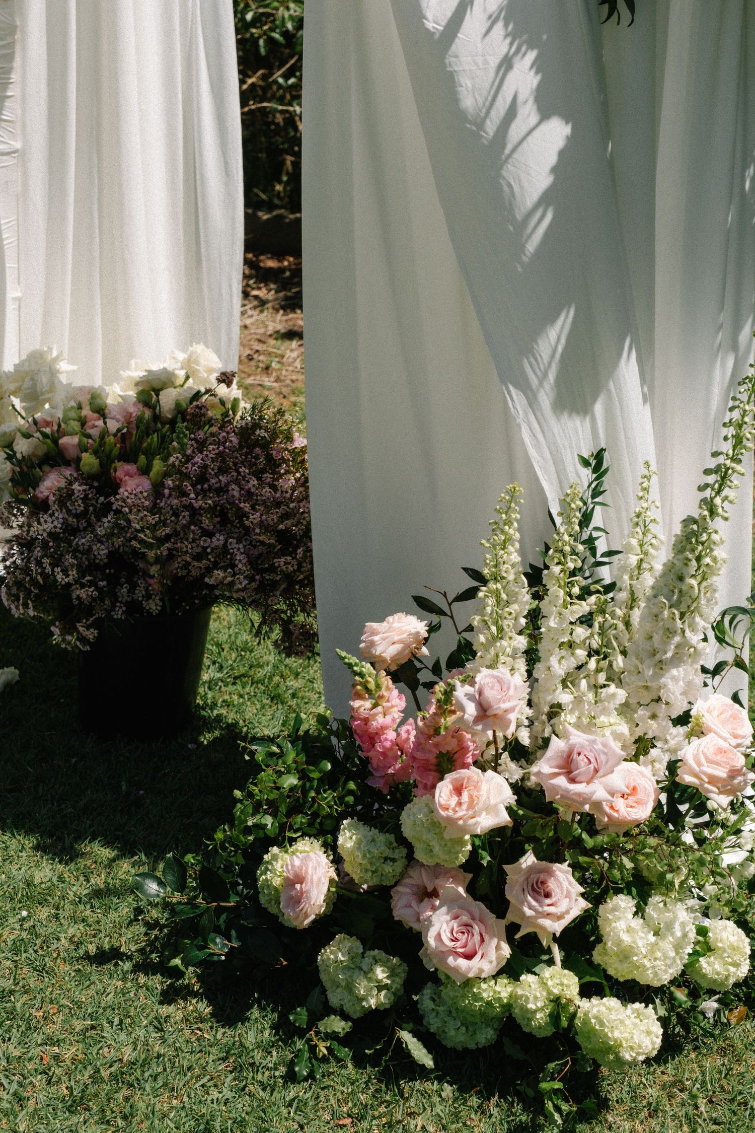 Pink roses and white hydrangeas create an elegant floral arrangement against flowing white curtains in outdoor setting.