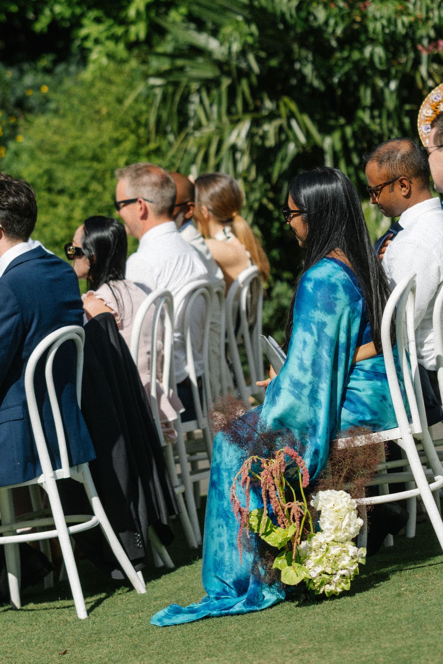 Wedding guests seated in white chairs on a green lawn with turquoise and dark colored attire at an outdoor ceremony.
