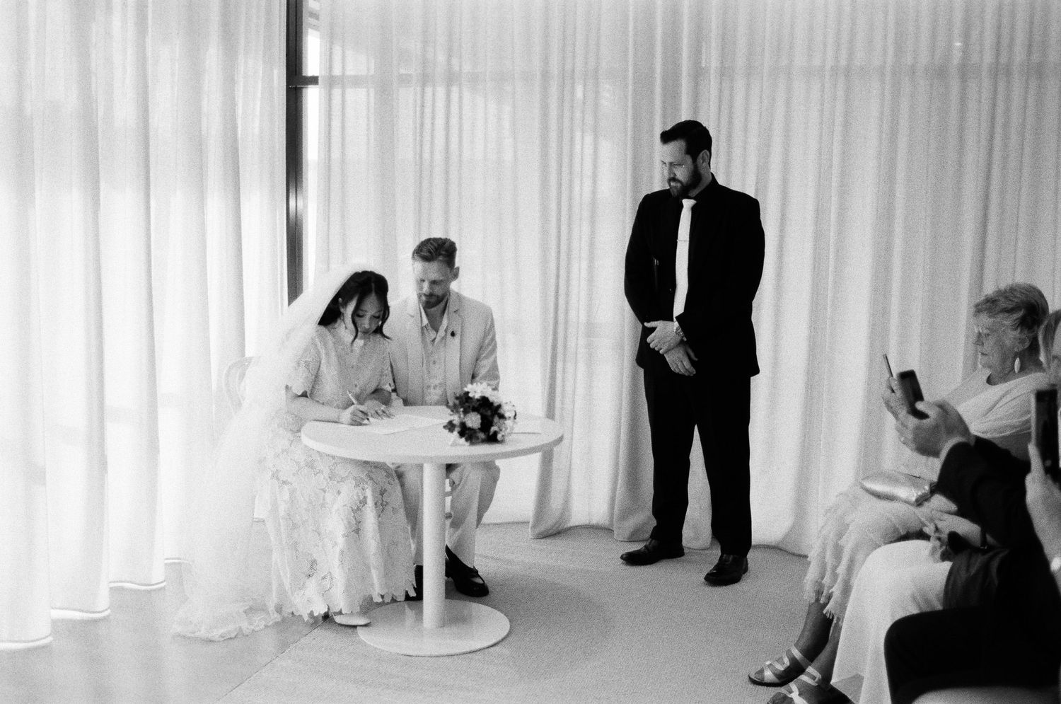 Black and white photo of a couple signing the marriage register at a small table while a groomsman and guests look on in a bright room.