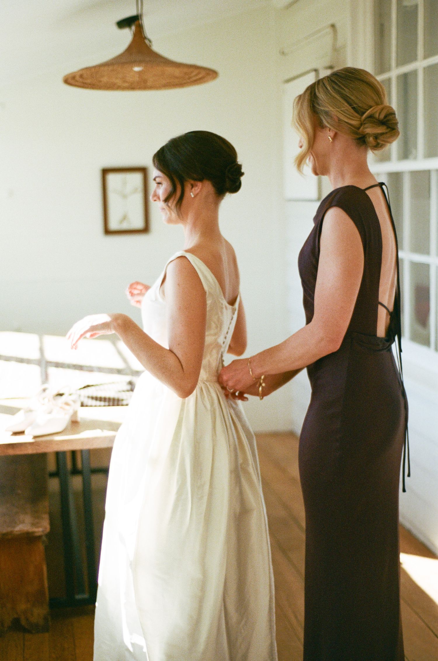 A bride in a white sleeveless wedding gown is helped with her dress by a woman in a dark open-back gown in a sunlit room.