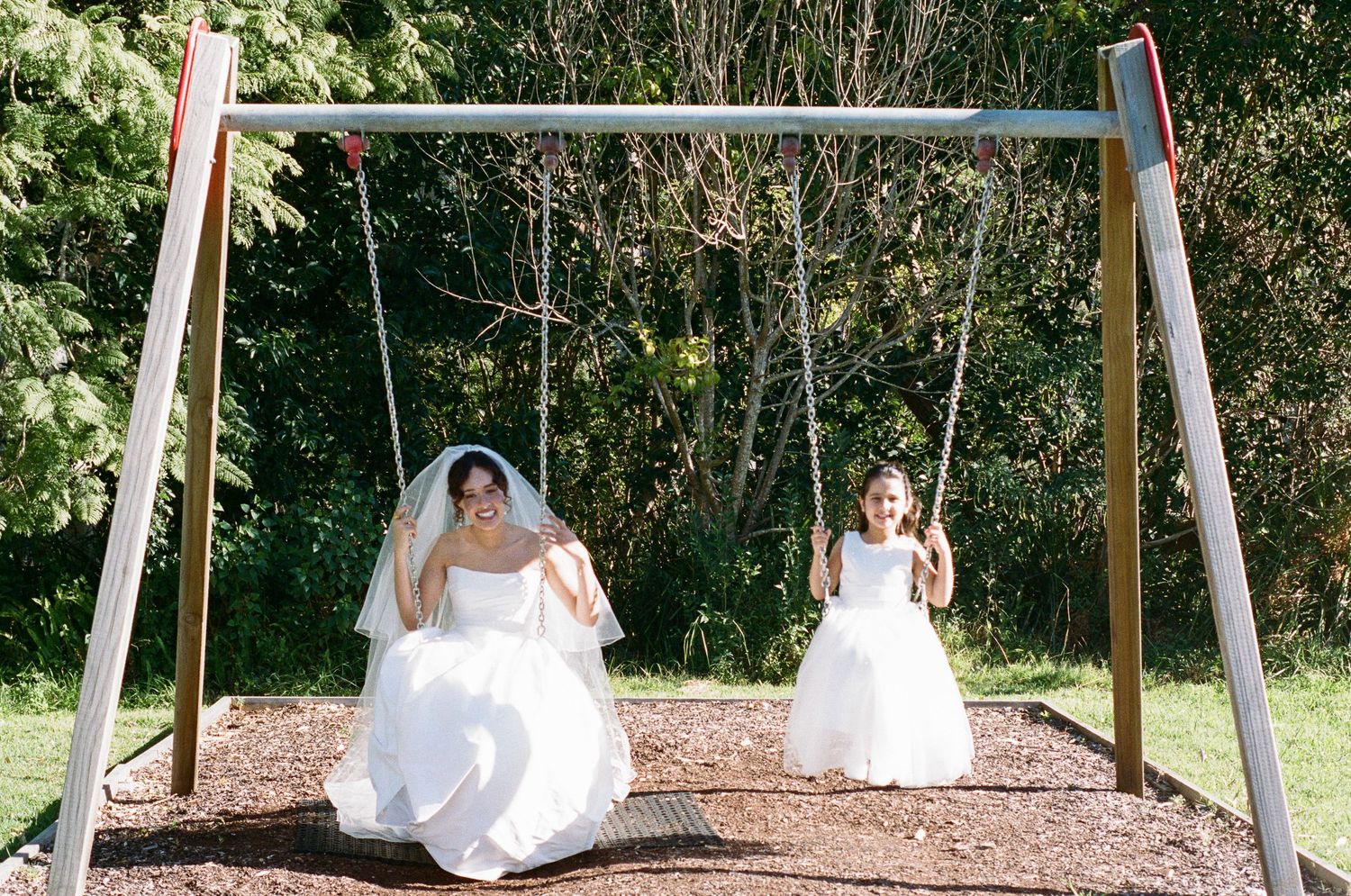 A bride in a white wedding dress and veil sits on a wooden swing beside a smiling flower girl in a white dress at a leafy park.