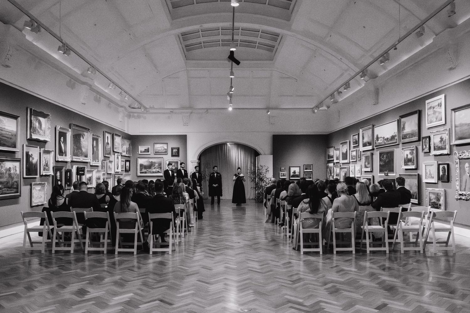 Art gallery interior with white walls displaying framed artwork and wooden chairs arranged in rows for viewing.