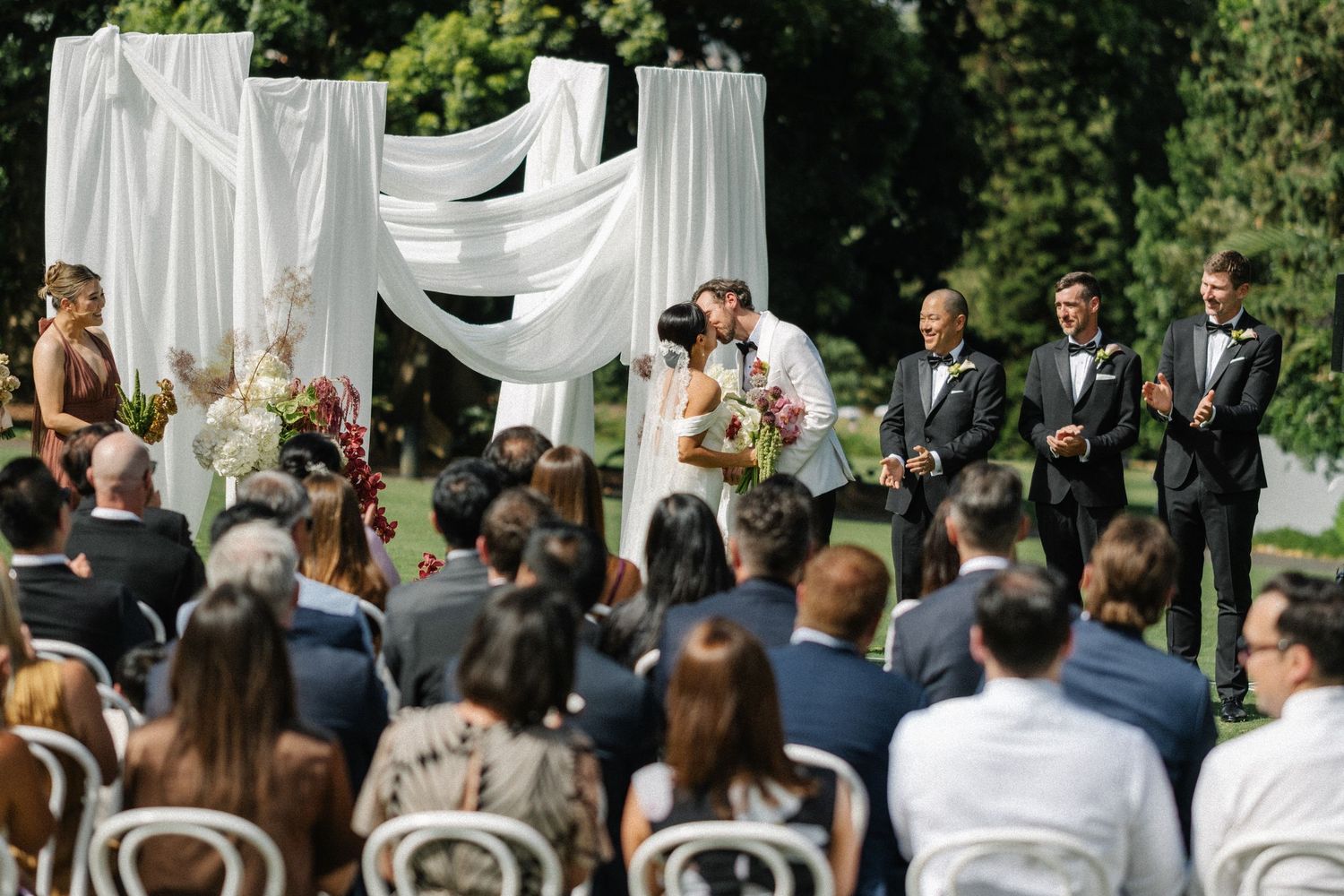 Wedding ceremony guests stand under white fabric draping outdoors surrounded by lush greenery and trees.