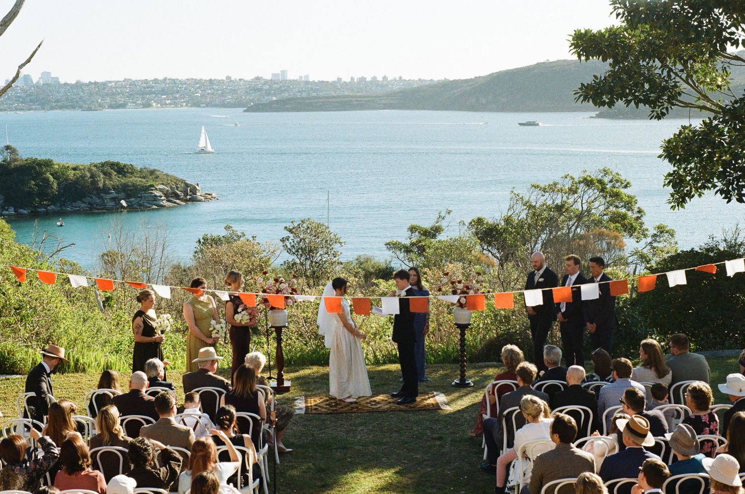 Outdoor waterfront wedding ceremony with guests seated on chairs as a bride and groom stand under orange bunting overlooking a sunny harbor.