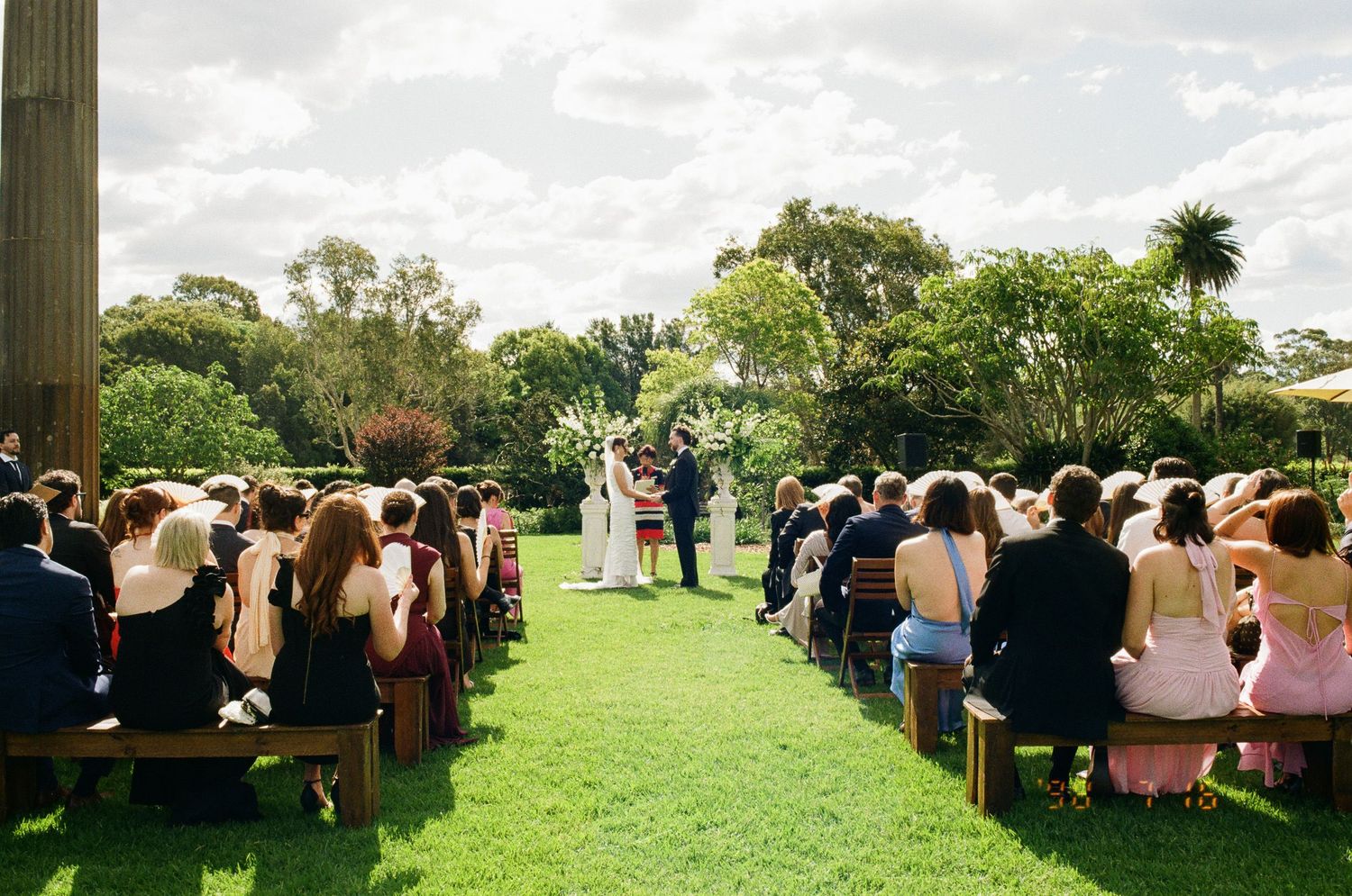 Wedding guests seated on wooden benches watch an outdoor garden ceremony as the couple stands at the altar on a sunny green lawn.