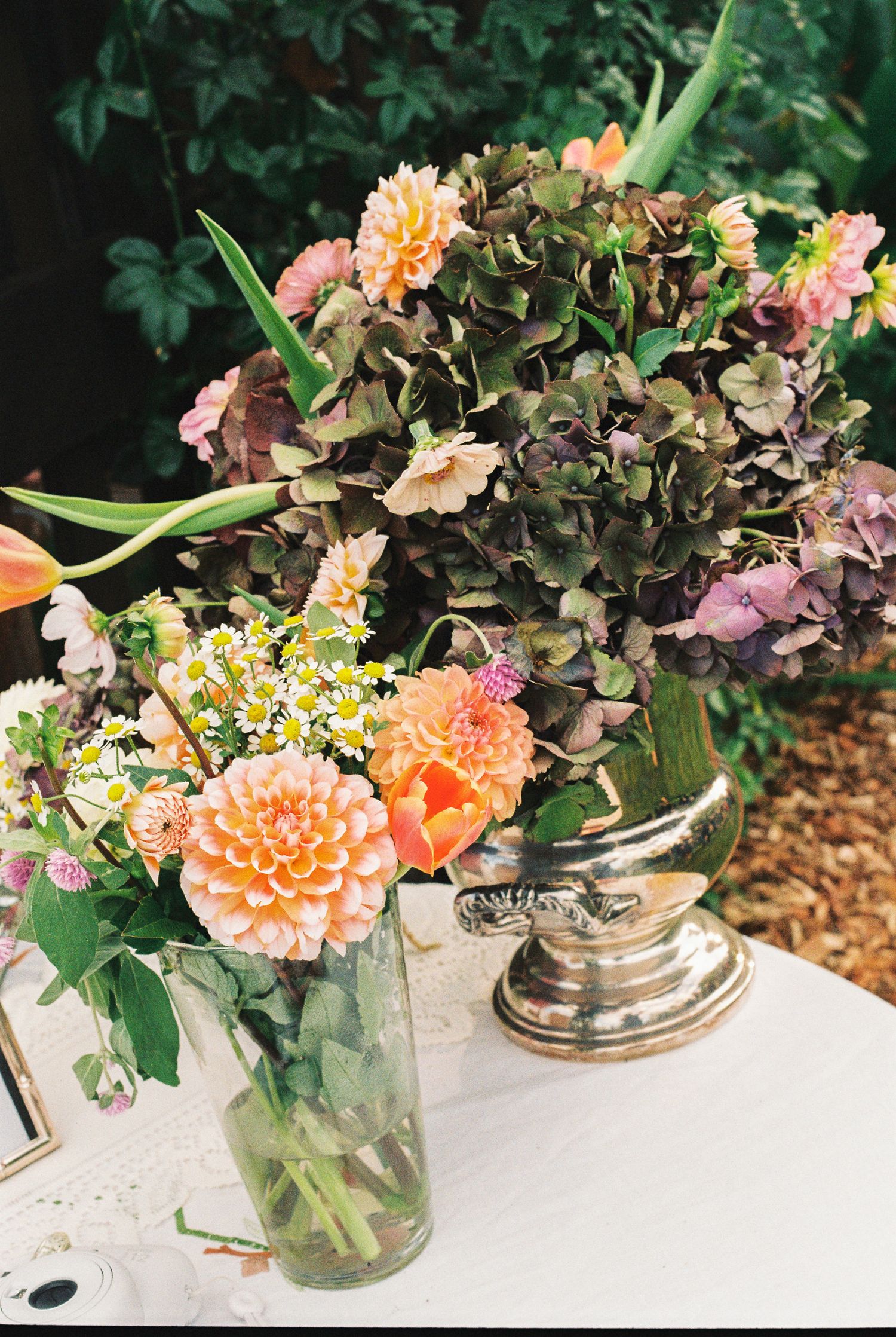 Colorful floral arrangement with peach dahlias, orange tulips, and dried green hydrangeas in a glass vase and silver urn on a white table.