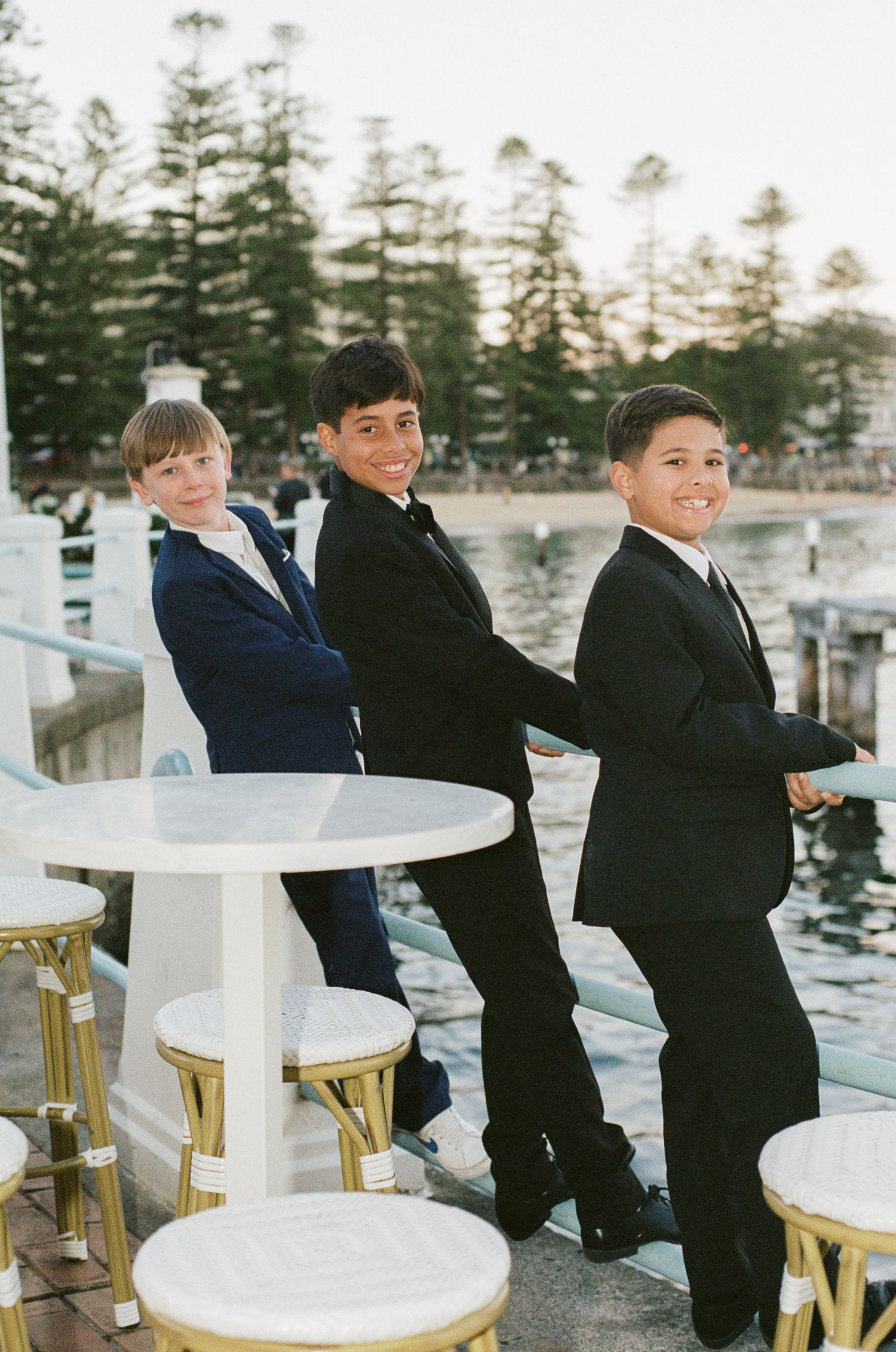 Three smiling boys in dark suits stand along a waterfront railing near white round cocktail tables and gold stools at an evening event.