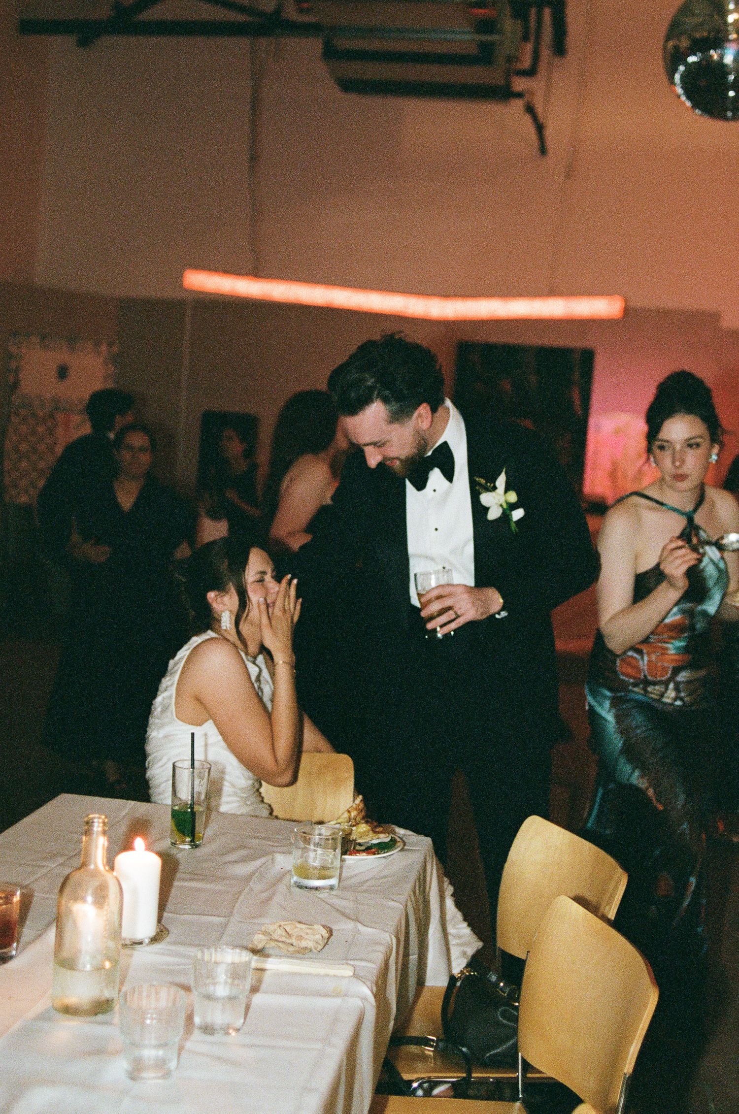 Groom in a black tuxedo and bow tie laughs beside a seated bride at a candlelit wedding reception table, with guests dancing behind them.