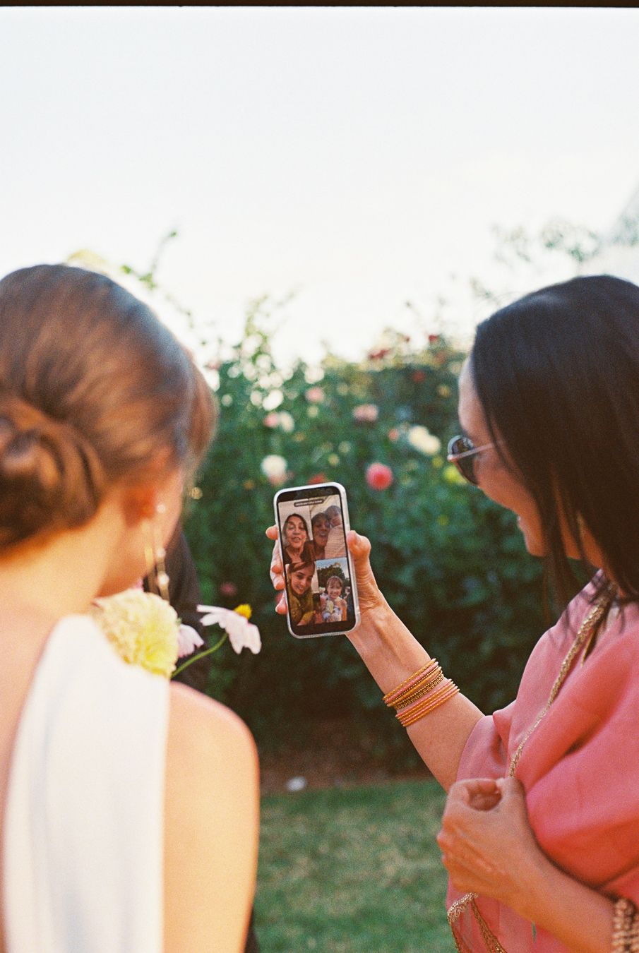 Wedding guest shows bride a FaceTime video call with family on smartphone at outdoor reception.