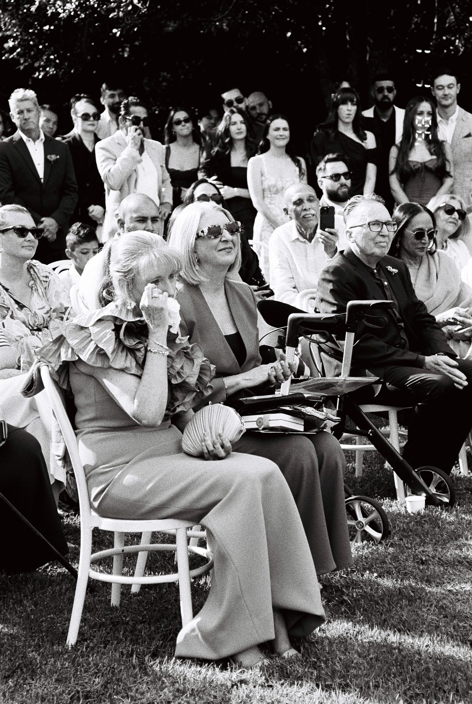 Emotional wedding guests seated outdoors on the grass, a woman in a ruffled dress wiping tears while the crowd watches the ceremony.