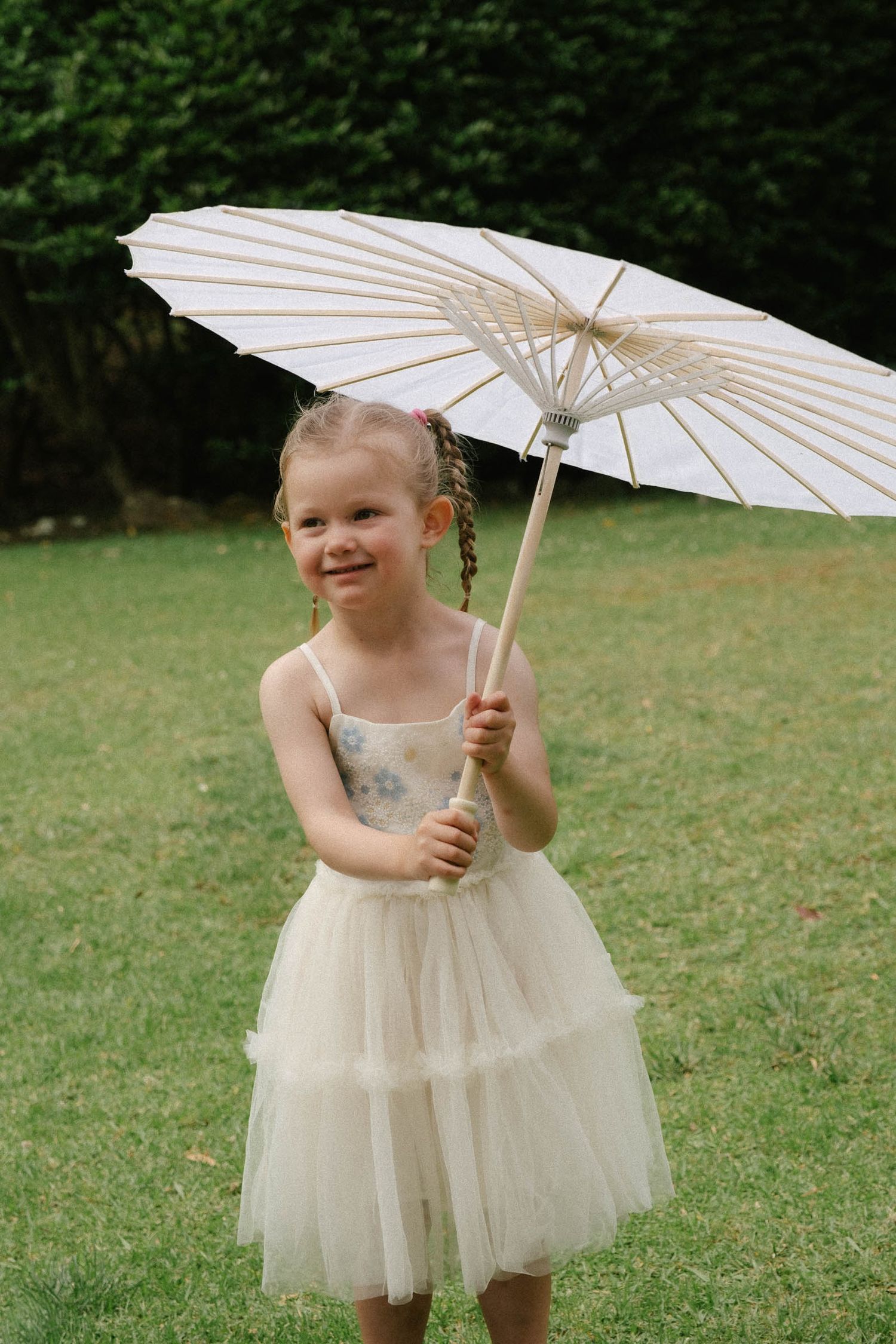 Small child in a cream tulle dress holding a white paper parasol on grass.