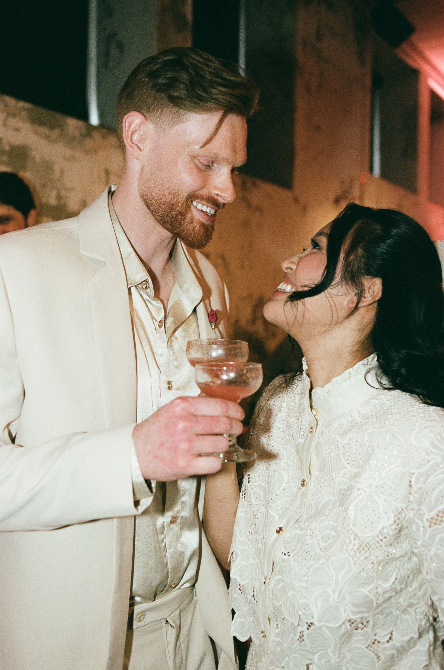 A smiling bearded man in a cream suit and a laughing woman in a white lace blouse share a toast with coupe glasses at a lively celebration.