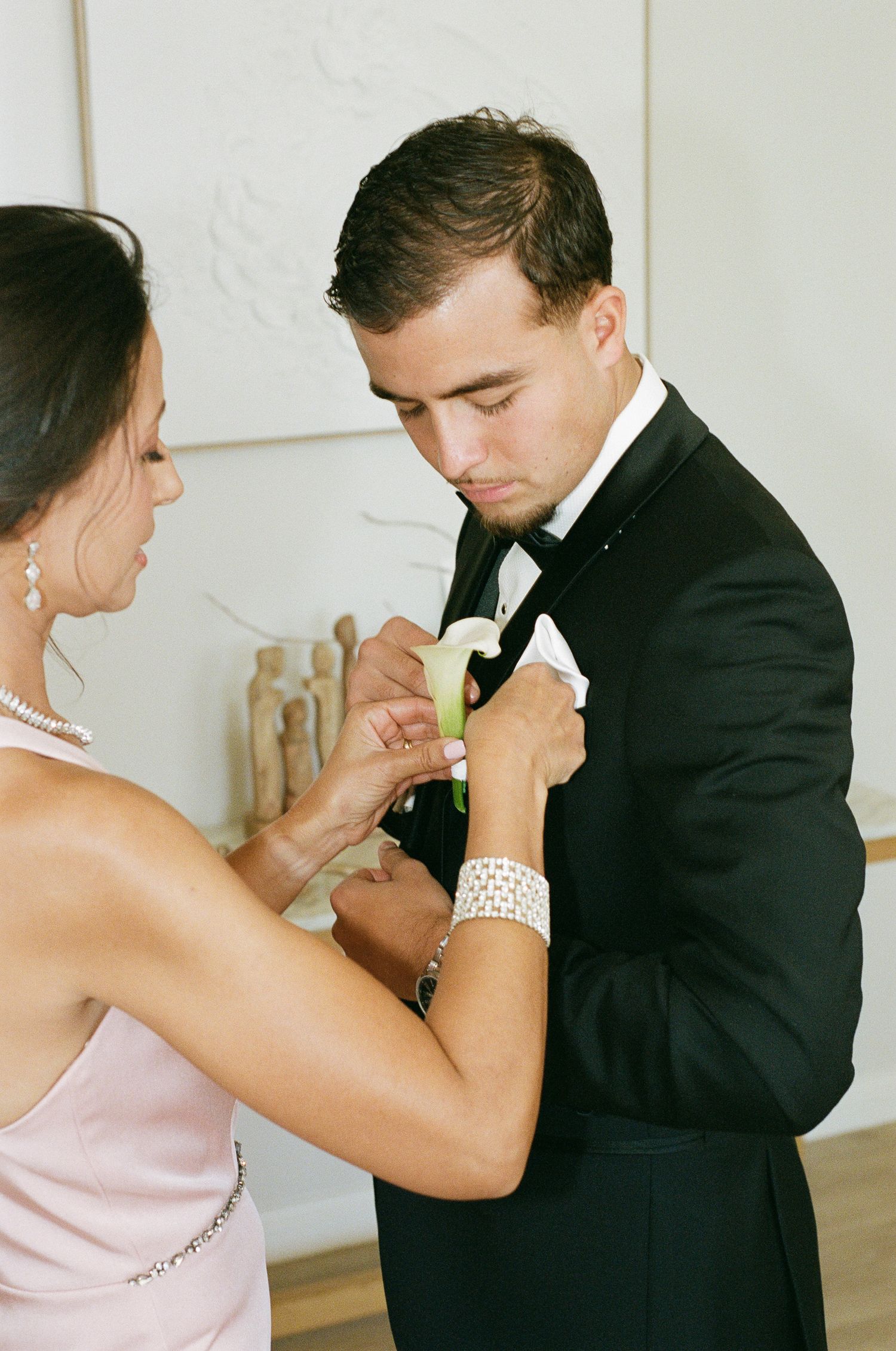 A woman in a blush pink dress pins a white calla lily boutonniere onto the black tuxedo lapel of a groom before the wedding.