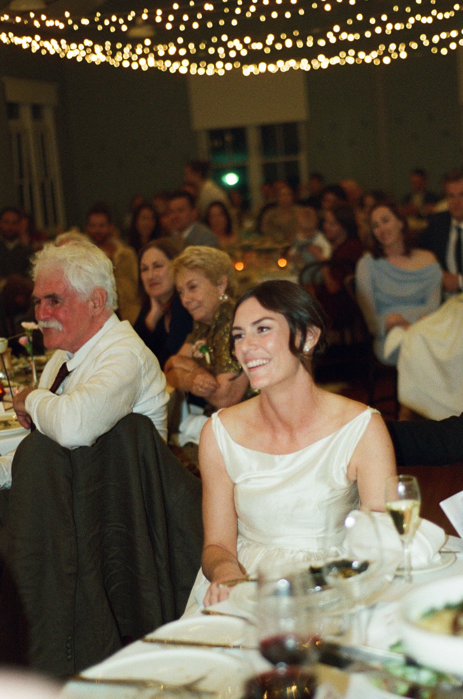 Smiling bride in a white dress sits at a reception table as guests laugh behind her under warm string lights.