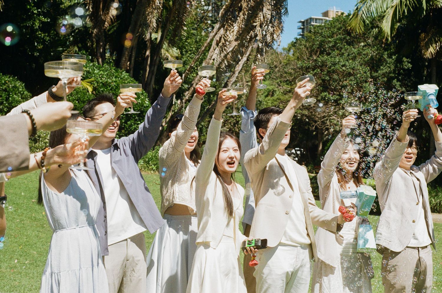 A group of friends in light, elegant outfits raise their glasses in a joyful outdoor toast under tall trees in a sunny park.